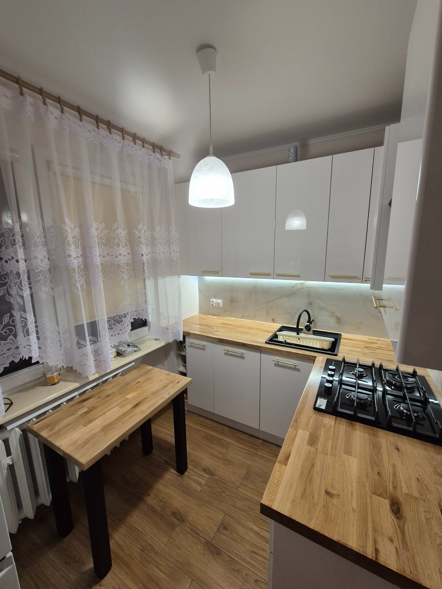 Jasna kuchnia z białymi szafkami, drewnianym blatem i stołem. Nad blatem wiszą lampy, a okno zdobią białe, wzorzyste firanki. Widoczna płyta gazowa i zlewozmywak.