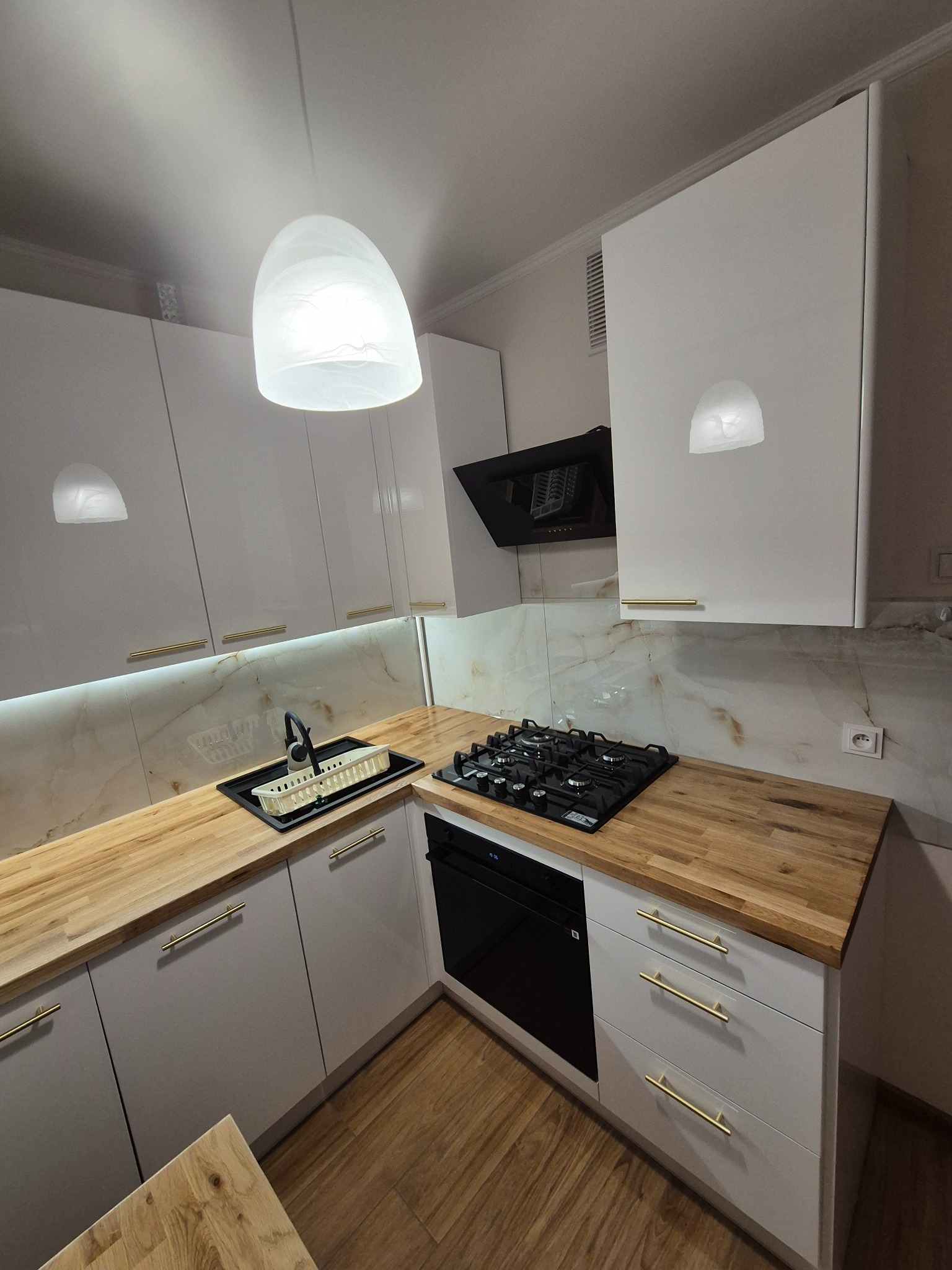 Nowoczesna kuchnia z białymi szafkami, drewnianym blatem i złotymi uchwytami. Oświetlenie LED pod szafkami górnymi. Widoczna płyta gazowa i okap.