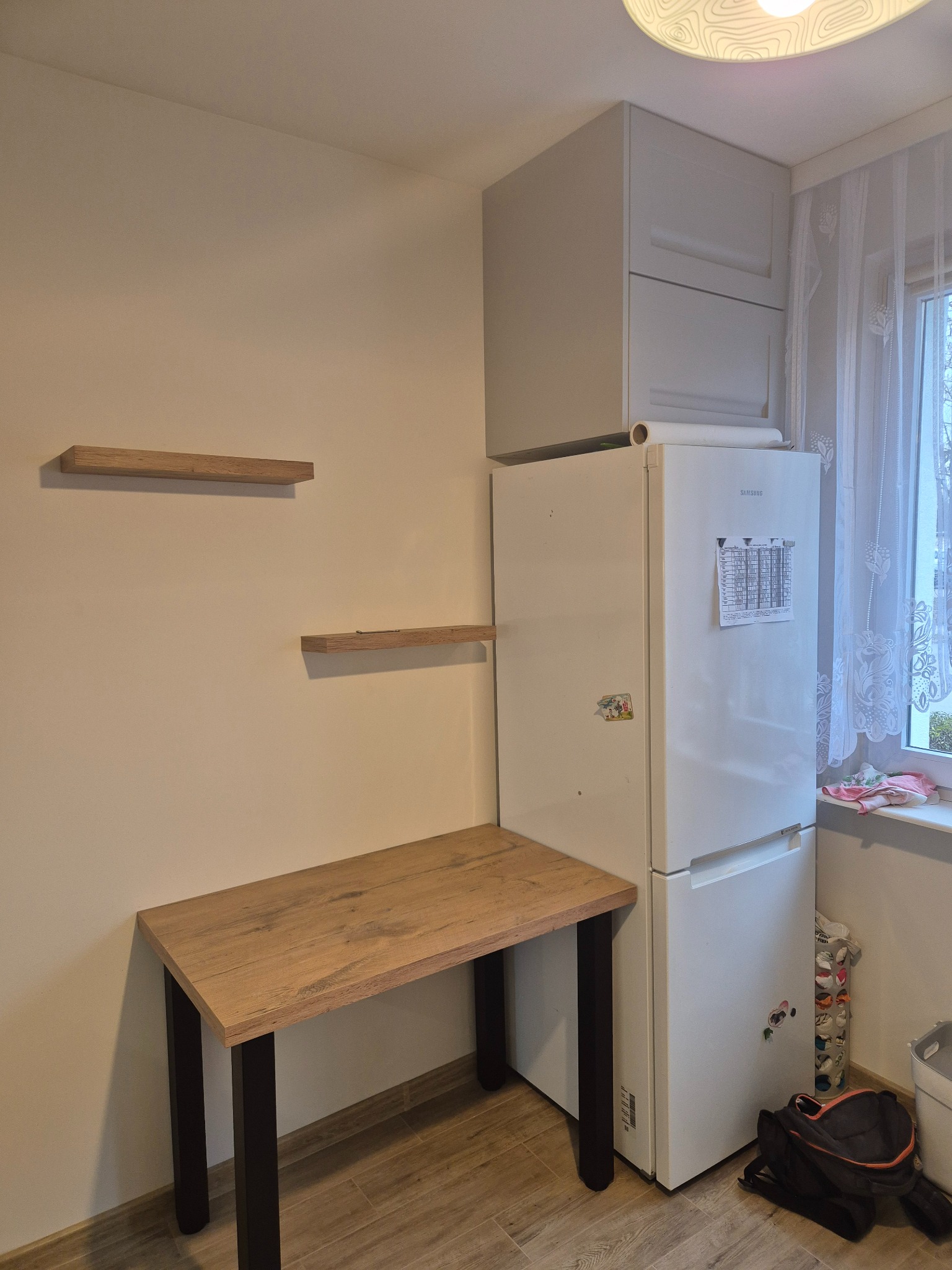 Kuchnia po montażu: drewniany blat biurka z czarnymi nogami, półki na ścianie, szafki wiszące nad lodówką. Widok na okno z firanką.