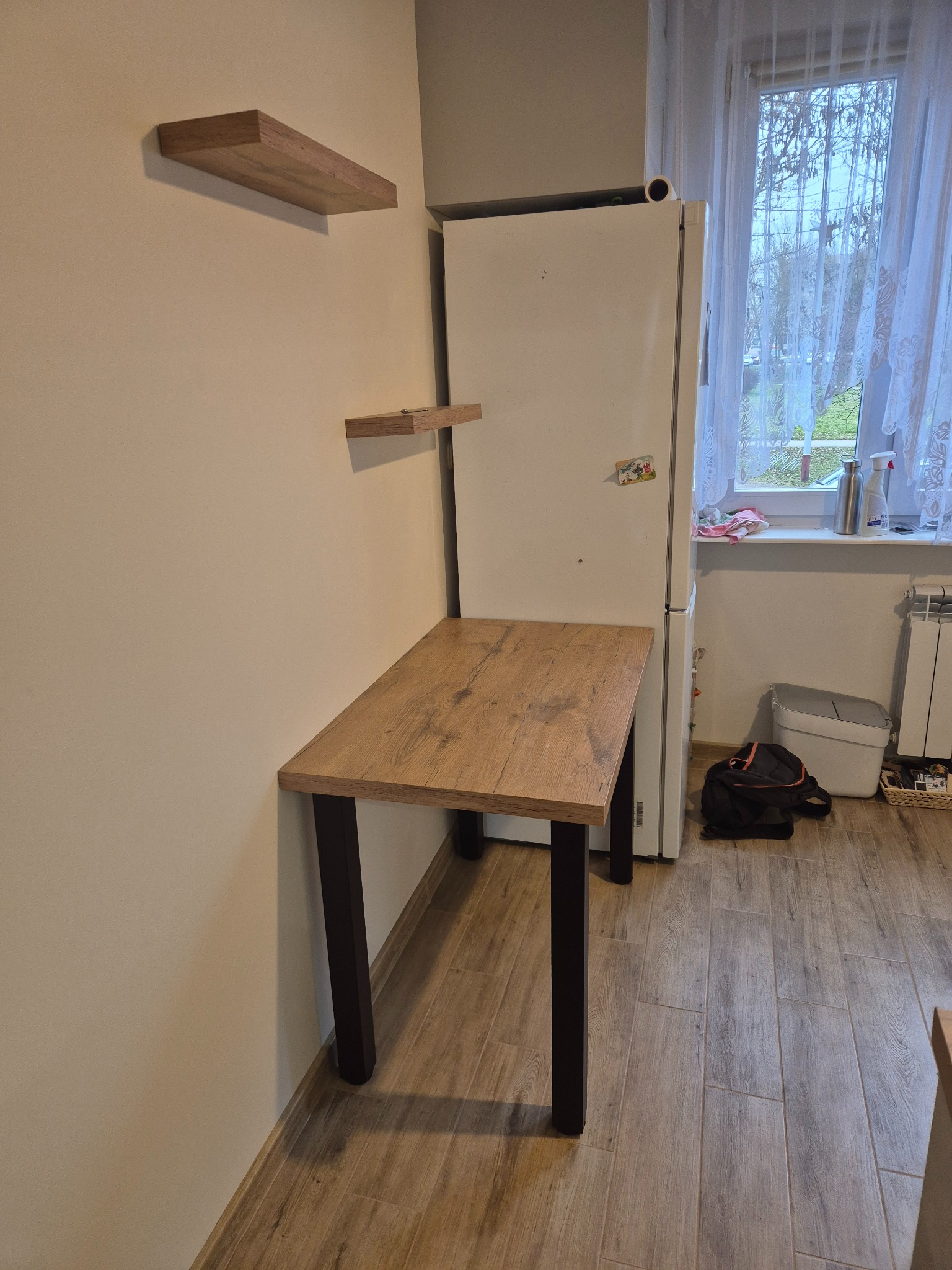 Zamontowany blat kuchenny z drewnianym dekorem, wsparty na czarnych nogach, przylegający do ściany. Nad blatem dwie półki w tym samym stylu. W tle lodówka i okno.