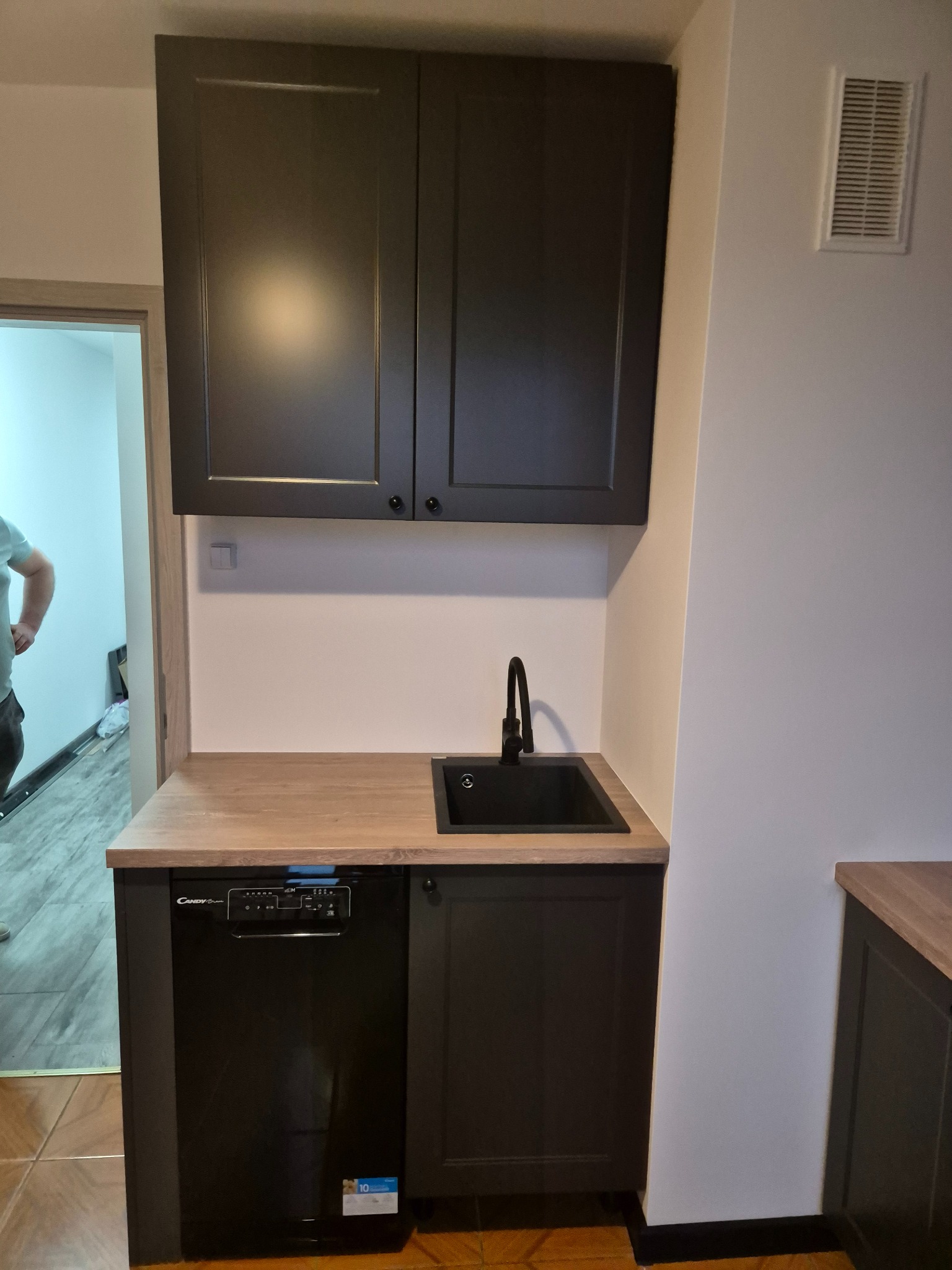 Ciemne szafki kuchenne z drewnianym blatem i czarnym zlewem, uzupełnione o zintegrowaną zmywarkę. Widok na fragment pomieszczenia z płytkami podłogowymi.