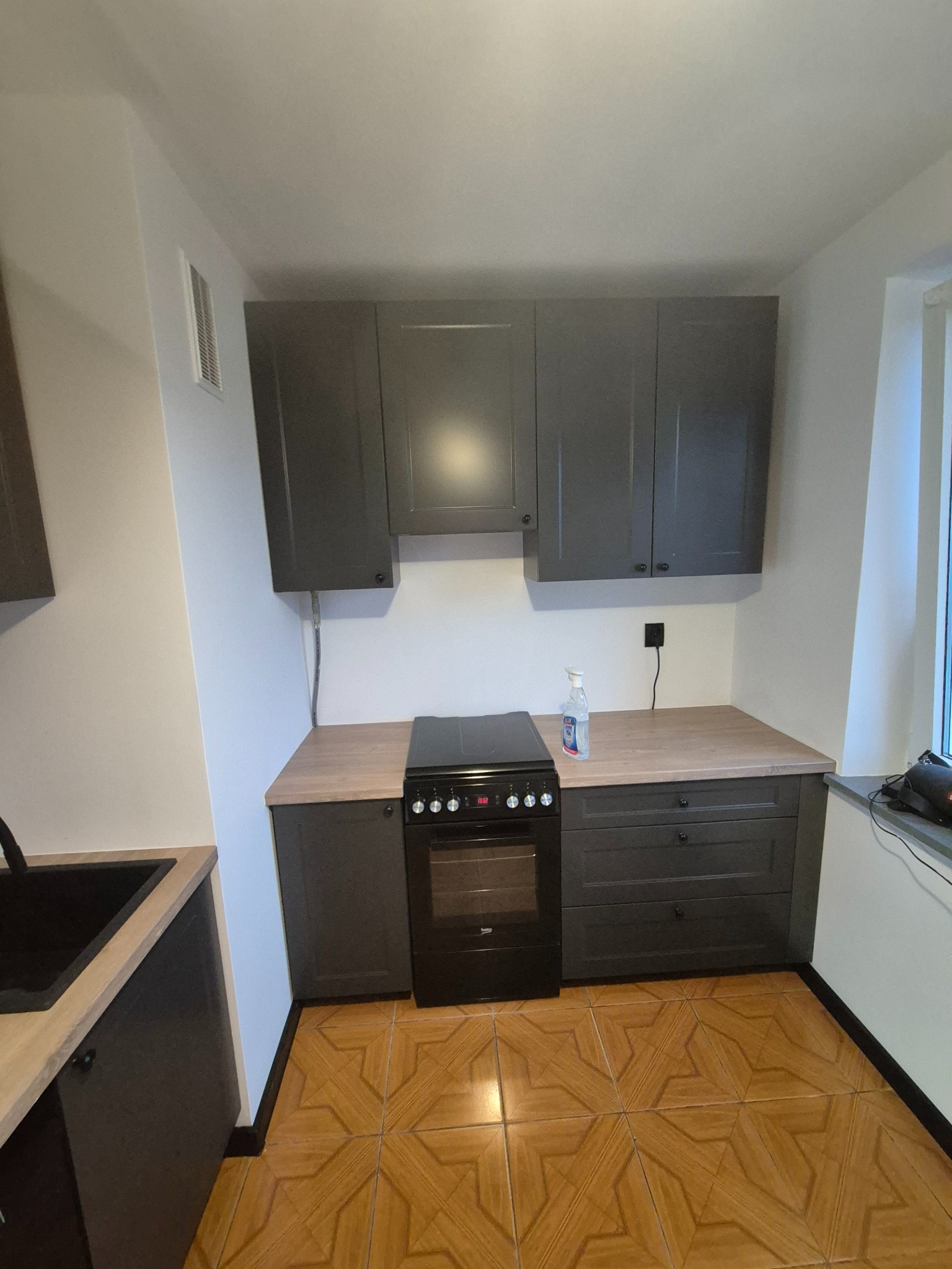 Szara zabudowa kuchenna z drewnianym blatem i płytą grzewczą, widok na fragment wnętrza z brązową podłogą. Ujęcie z perspektywy podłogi.