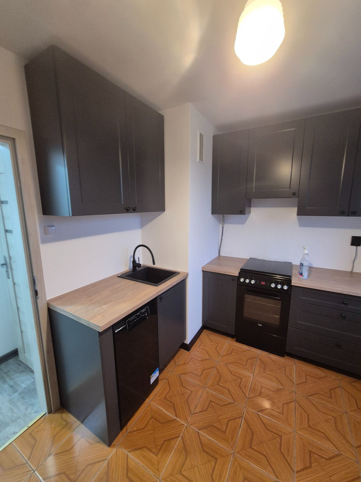 Szara zabudowa kuchenna z drewnianym blatem, czarnym zlewem i płytą. Uwagę zwracają brązowe płytki podłogowe w geometryczny wzór. Widok na fragment kuchni po remoncie.