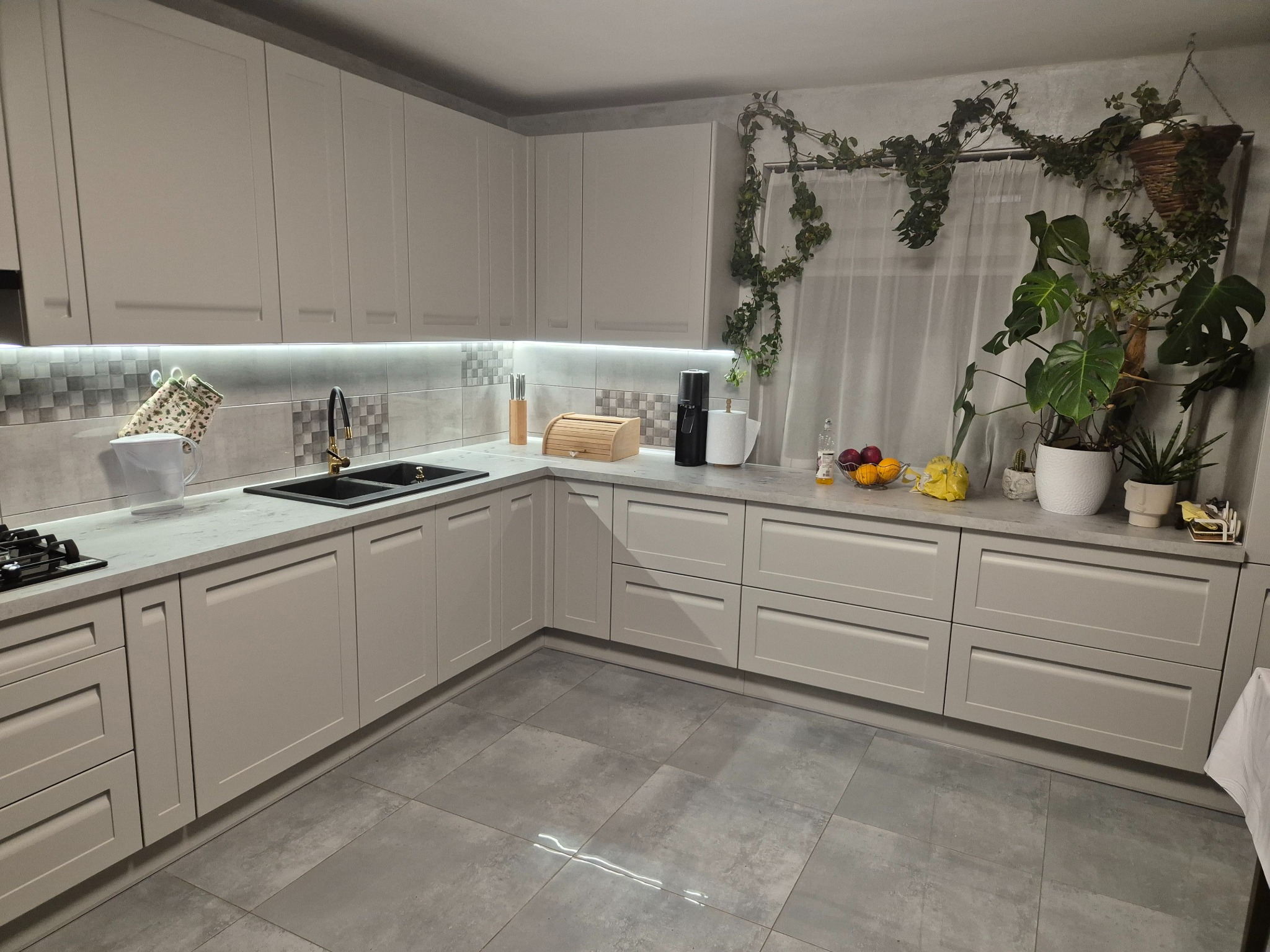 Jasna kuchnia z szarymi frontami, blatem i płytkami. Nad blatem wiszące szafki, a na nim akcesoria kuchenne i rośliny doniczkowe. Ujęcie z perspektywy podłogi.