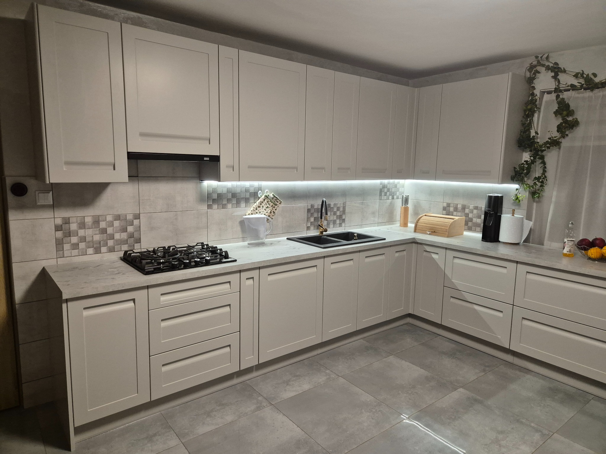 Jasna kuchnia w stylu skandynawskim z szarymi szafkami, płytkami i blatem. Nad blatem zamontowane oświetlenie LED. Na blacie widoczne akcesoria kuchenne.