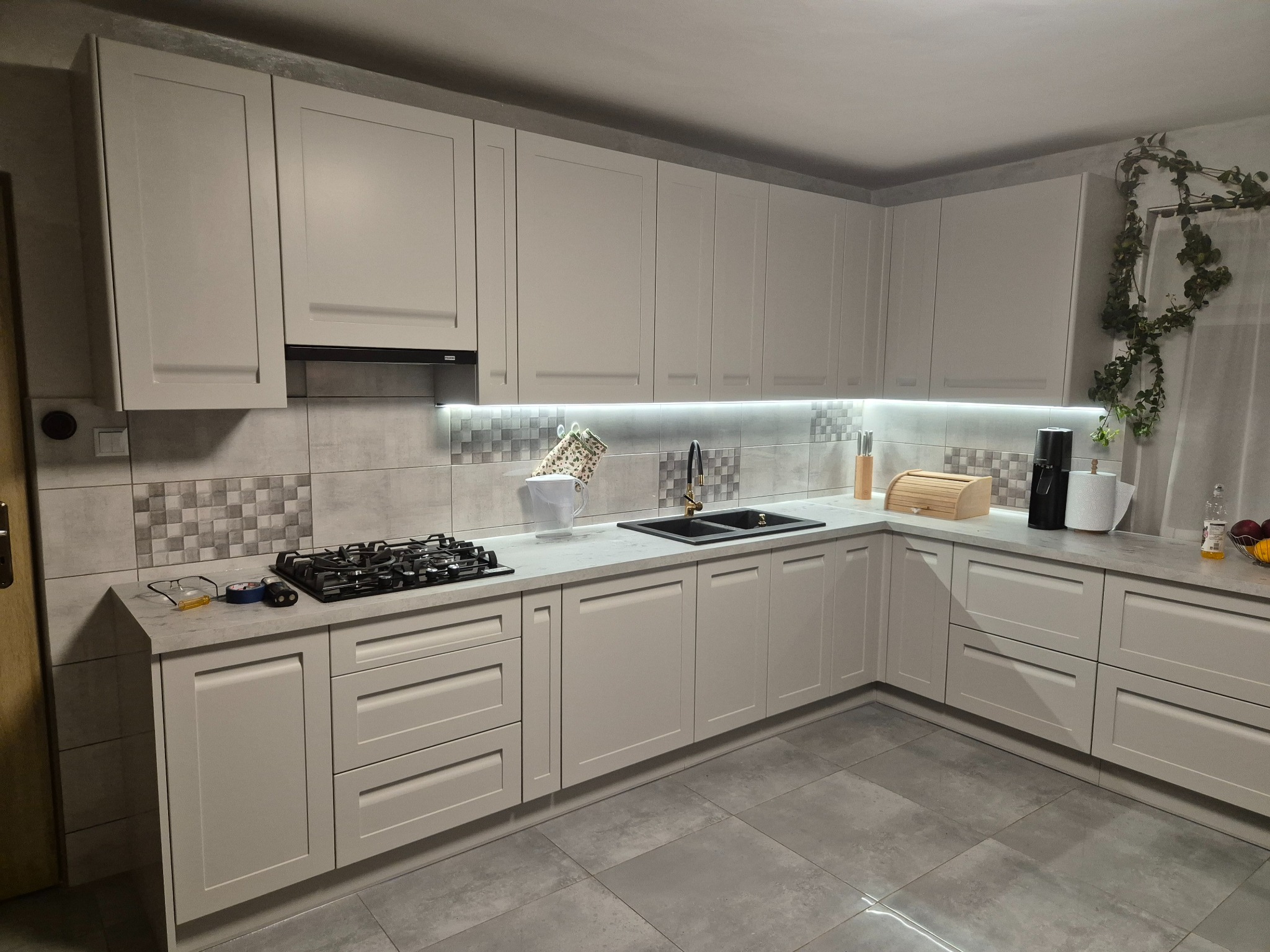 Jasna, narożna zabudowa kuchenna z szarymi frontami, czarnym zlewem i płytą gazową. Uwagę zwraca dekoracja z roślin pnących po prawej stronie.