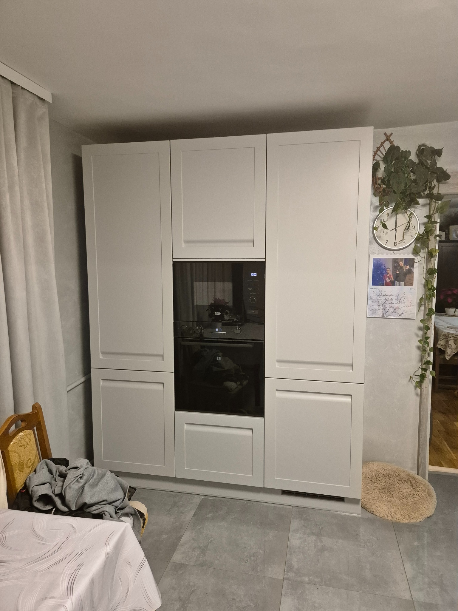 Biała zabudowa kuchenna z piekarnikiem i szafkami, widok z przodu, szare płytki podłogowe, dekoracje roślinne na ścianie, krzesło i stół z boku.