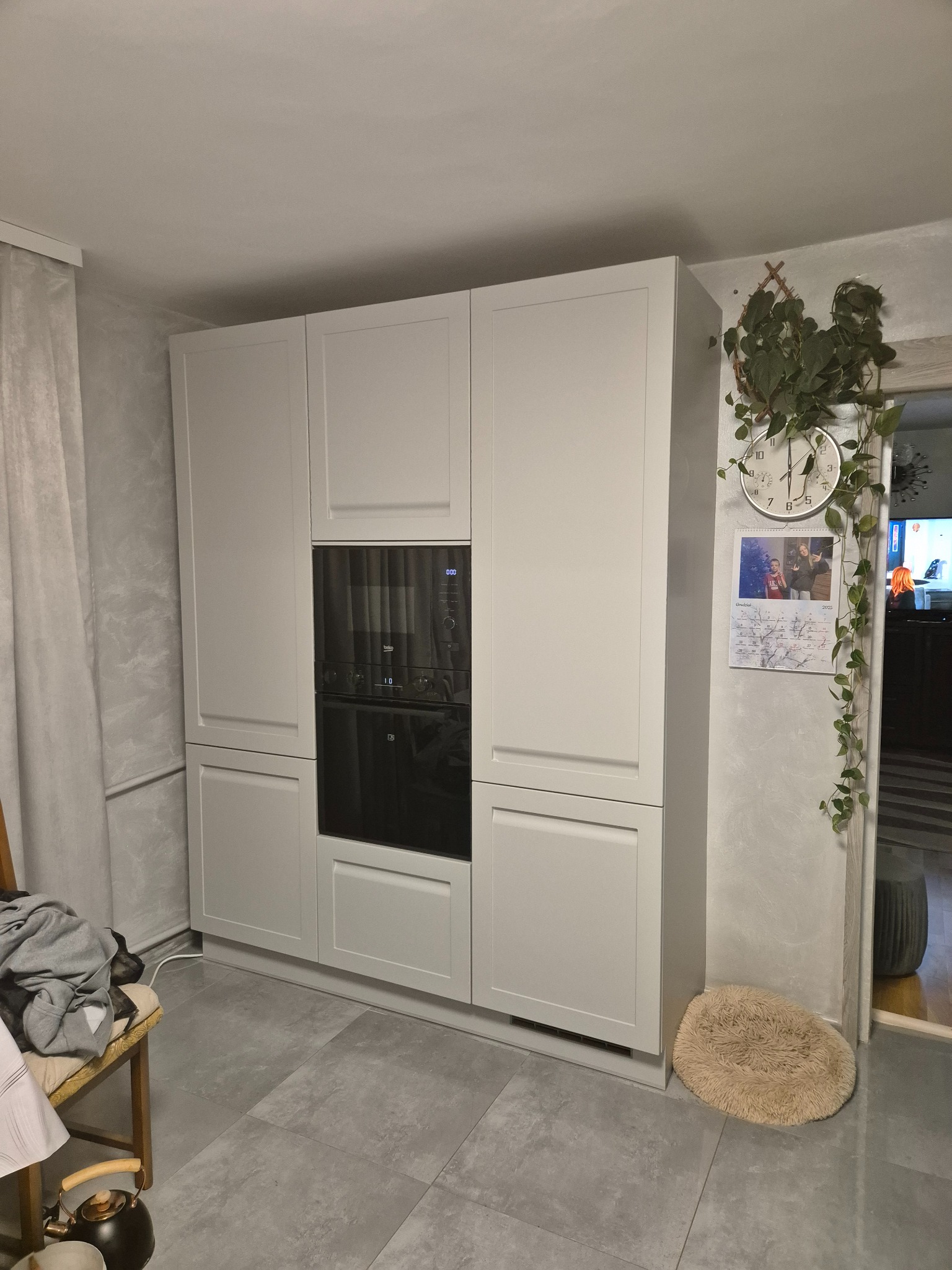 Biała zabudowa kuchenna z piekarnikiem w nowoczesnym wnętrzu. Na ścianie zegar i kalendarz, obok wejścia pnąca roślina. Szare płytki podłogowe.