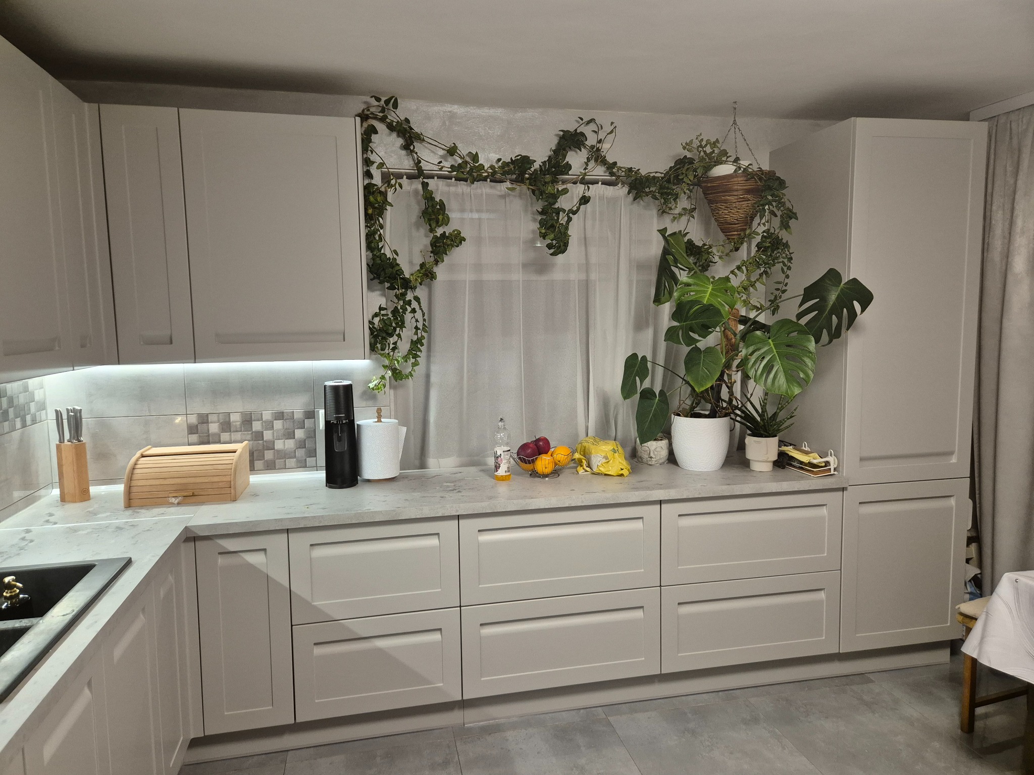 Jasna kuchnia z szarymi szafkami, blatem i roślinami pnącymi się nad oknem. Na blacie owoce, chlebak i akcesoria kuchenne. Minimalistyczny wystrój.