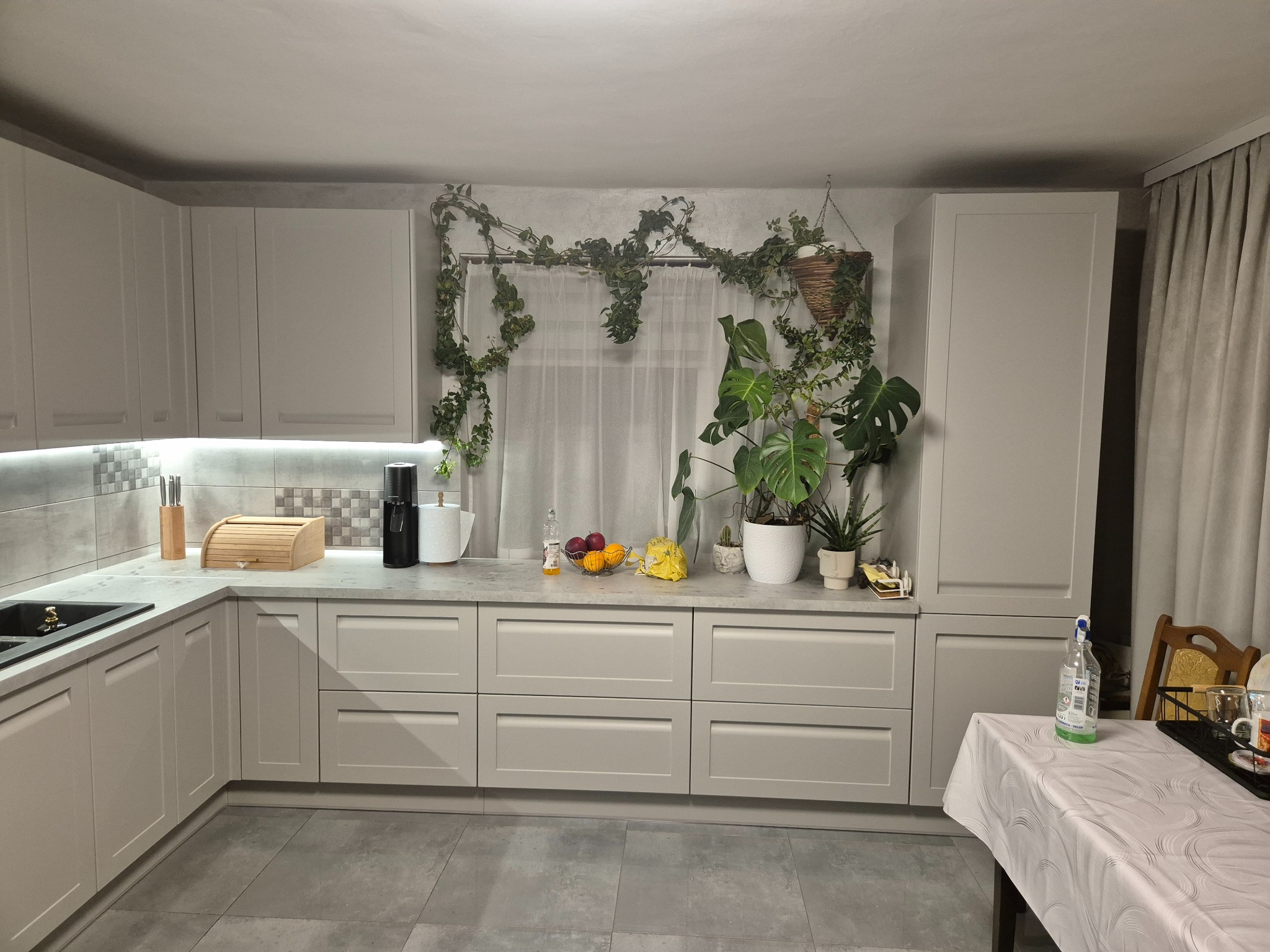 Jasna kuchnia z szarymi frontami szafek, blatem i roślinami pnącymi się nad oknem. Na blacie owoce i akcesoria kuchenne. Minimalistyczny wystrój.