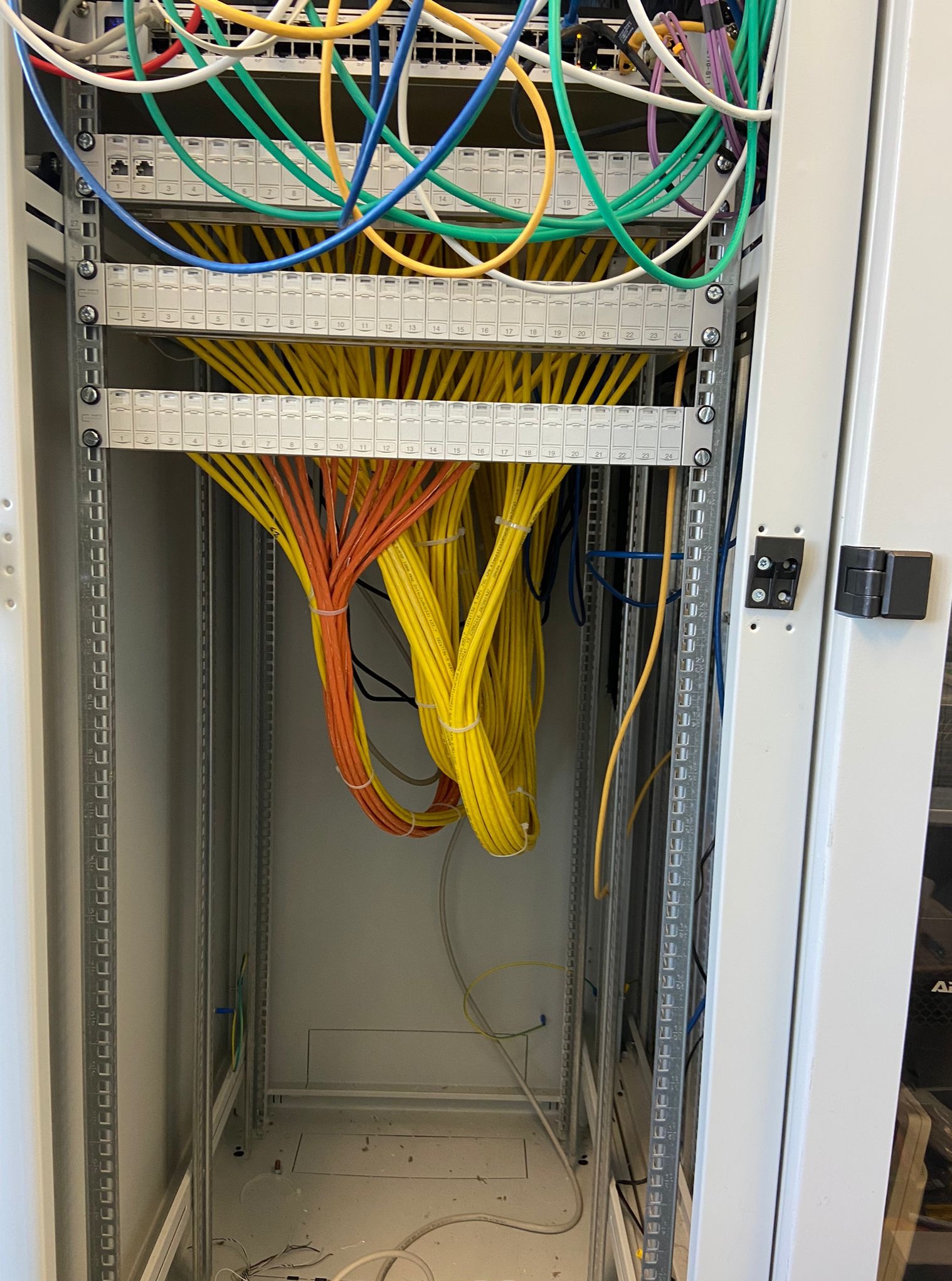 Szafa serwerowa z uporządkowanymi kablami sieciowymi w kolorach: żółtym, pomarańczowym, zielonym i niebieskim. Widoczne złącza i metalowa konstrukcja szafy.