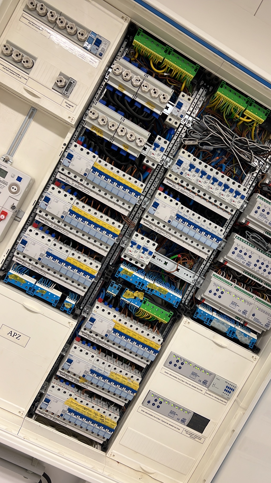 Rozdzielnica elektryczna z licznymi bezpiecznikami, przekaźnikami i kablami. Widoczne oznaczenia APZ i elementy sterujące. Ujęcie pod kątem.