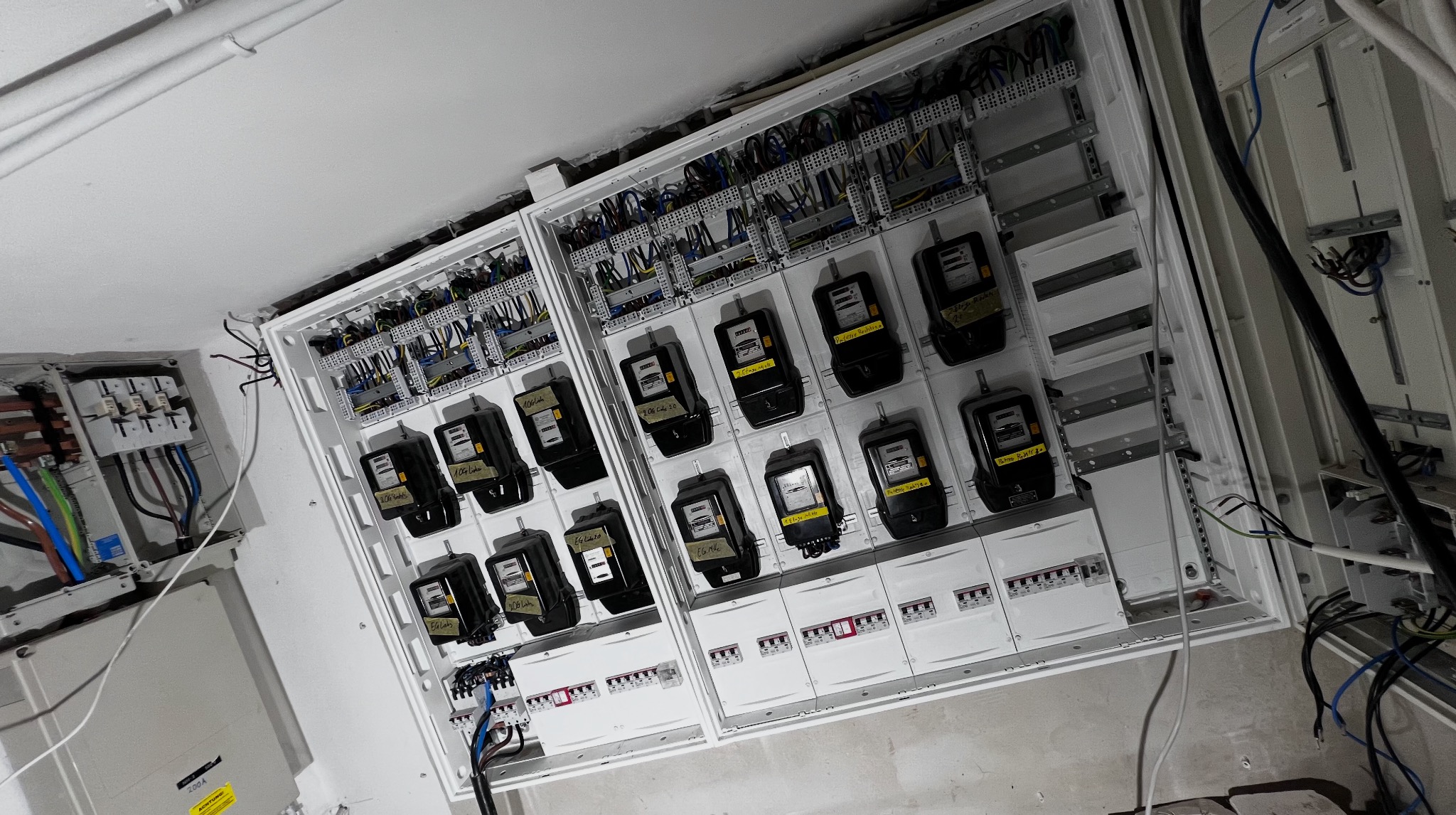 Rozdzielnia elektryczna z licznikami i okablowaniem w białej obudowie, widoczne bezpieczniki i oznaczenia na licznikach. Instalacja wewnątrz budynku.