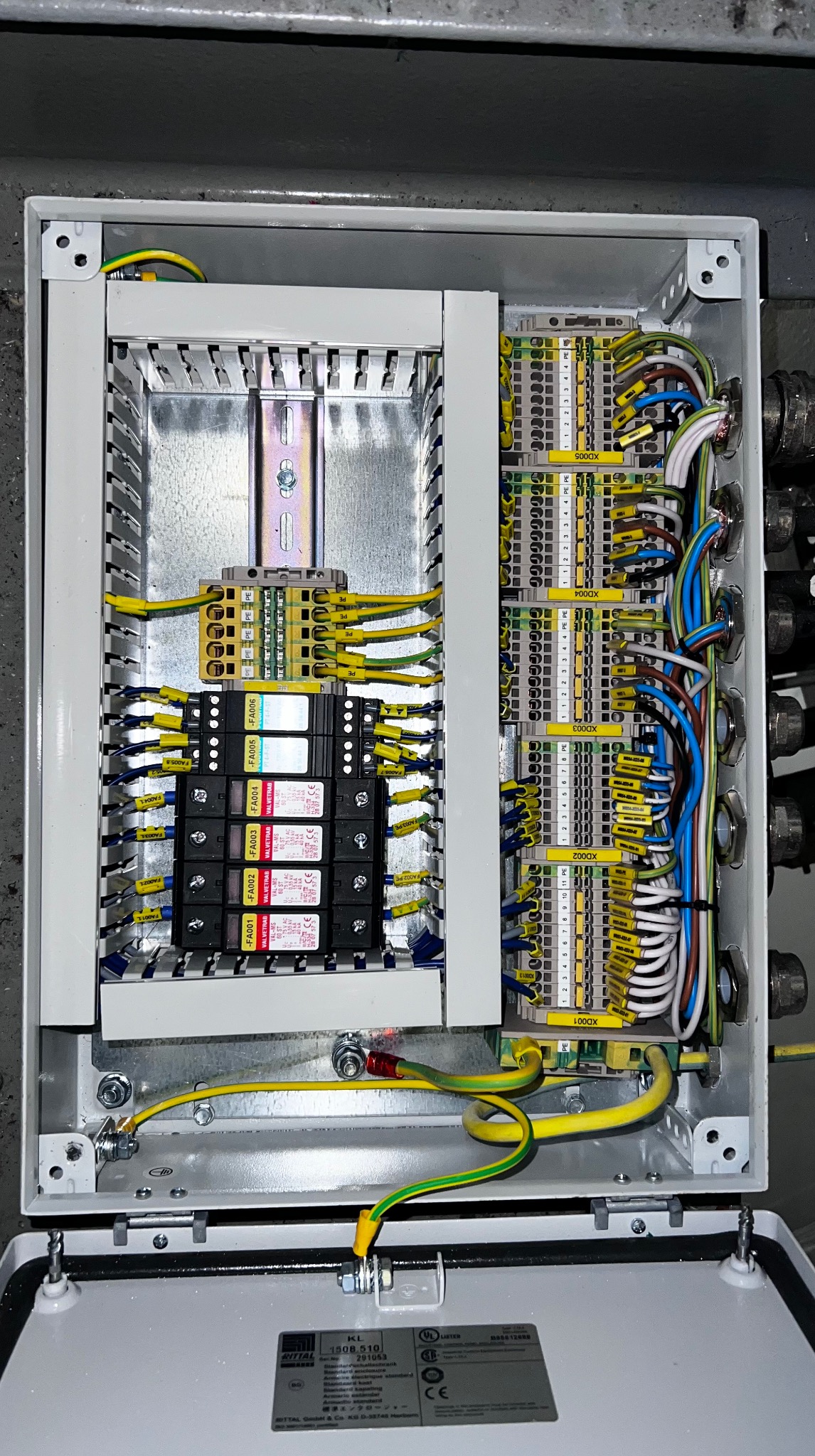 Otwarta skrzynka elektryczna z licznymi przewodami i złączkami, widoczne elementy zabezpieczeń i uziemienia. Precyzyjna instalacja w szarej obudowie.