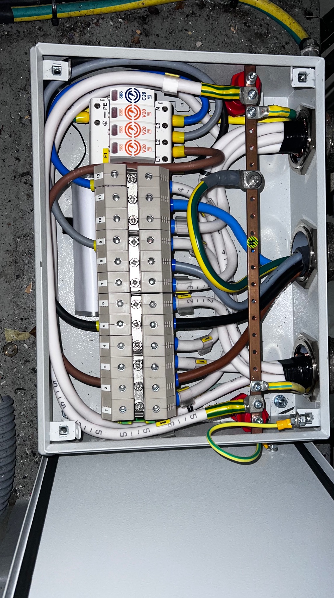 Otwarta skrzynka elektryczna z precyzyjnie ułożonymi przewodami, listwami zaciskowymi i zabezpieczeniami przepięciowymi. Widoczne oznaczenia przewodów i elementów.