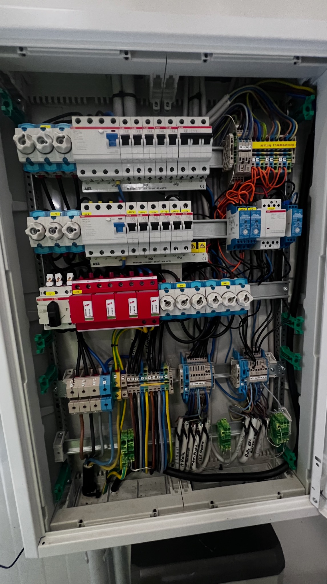 Otwarta skrzynka elektryczna z bezpiecznikami, przewodami i elementami montażowymi, widoczne różne kolory przewodów i oznaczenia na elementach.