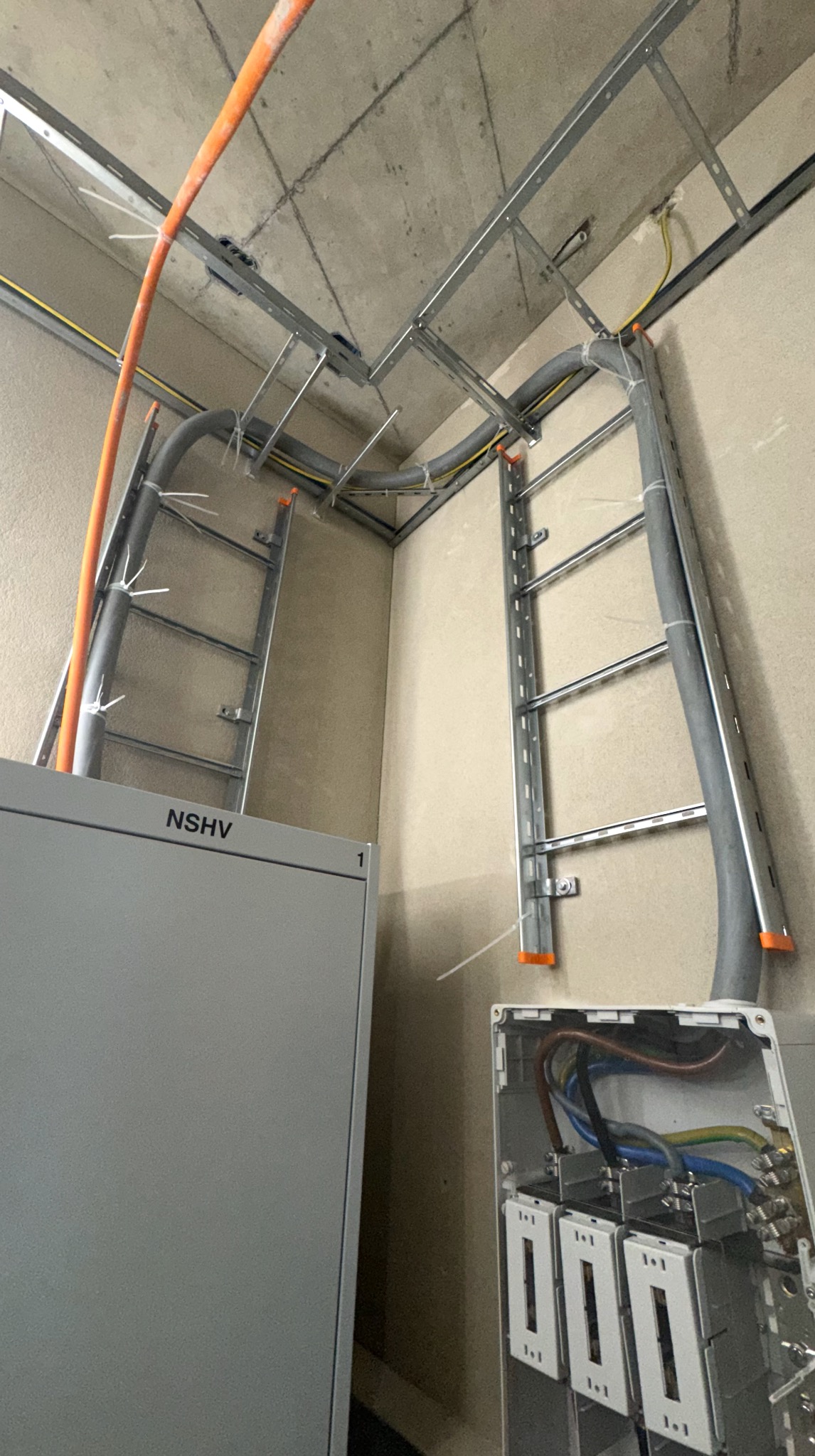 Instalacja elektryczna w surowym wnętrzu: koryta kablowe pod sufitem, szare rury z kablami, skrzynka NSHV i otwarta puszka z widocznymi przewodami.