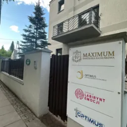 Tablica informacyjna z logo 'MAXIMUM Kancelaria Rachunkowa' przed budynkiem. Widoczne również logo innych firm. Ujęcie z poziomu chodnika, w tle iglaste drzewo.