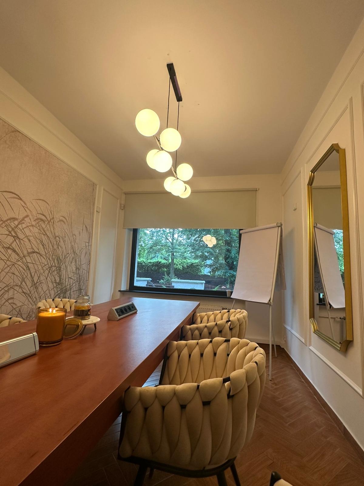 Sala konferencyjna z długim stołem, krzesłami, oknem z widokiem na zieleń i tablicą suchościeralną. Nowoczesne oświetlenie i dekoracje tworzą profesjonalną atmosferę.