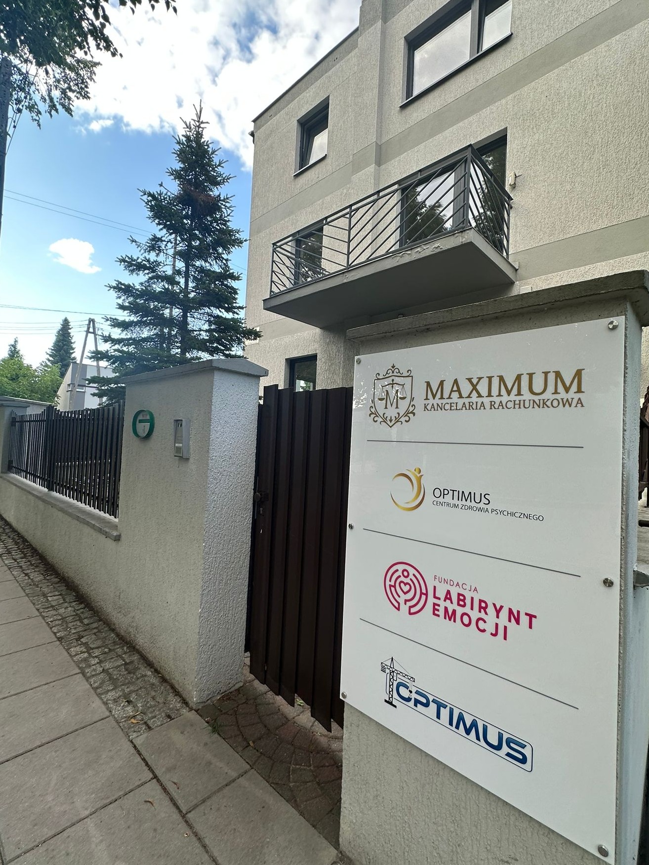 Tablica informacyjna z logo 'MAXIMUM Kancelaria Rachunkowa' przed budynkiem. Widoczne również logo innych firm. Ujęcie z poziomu chodnika, w tle iglaste drzewo.