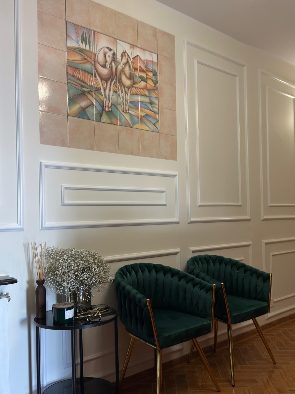 Eleganckie wnętrze: Dwa zielone krzesła z weluru na złotych nogach, stolik z kwiatami i dekoracyjna mozaika z motywem koni na ścianie z białymi panelami.
