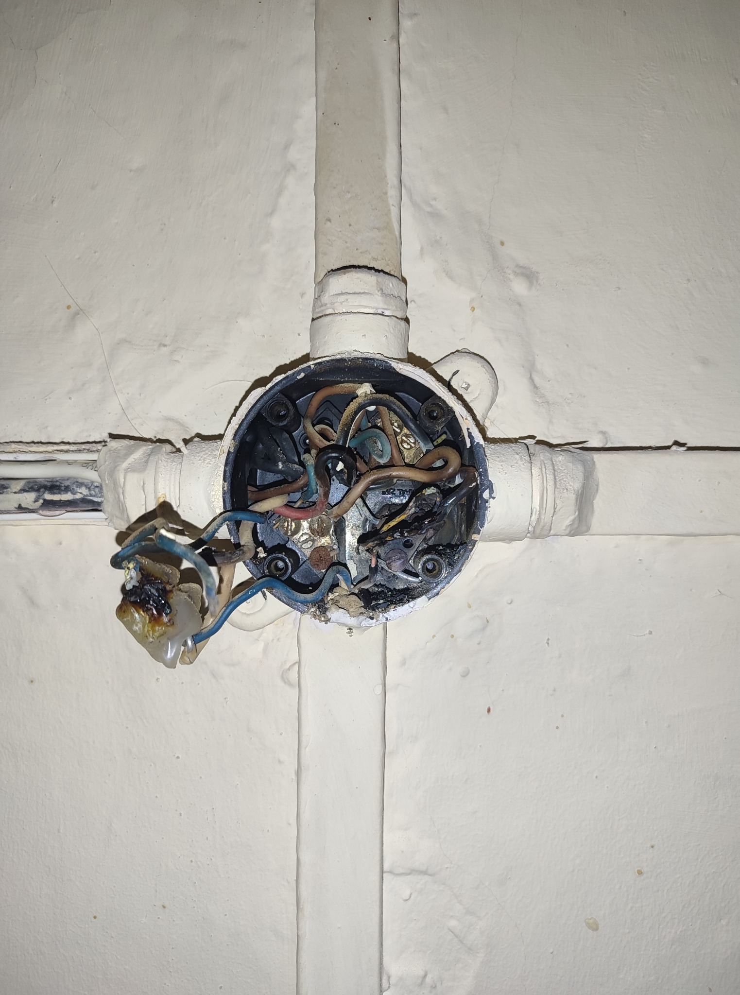 Uszkodzona, wypalona puszka elektryczna z widocznymi stopionymi przewodami na tle ściany. Awaria instalacji elektrycznej.