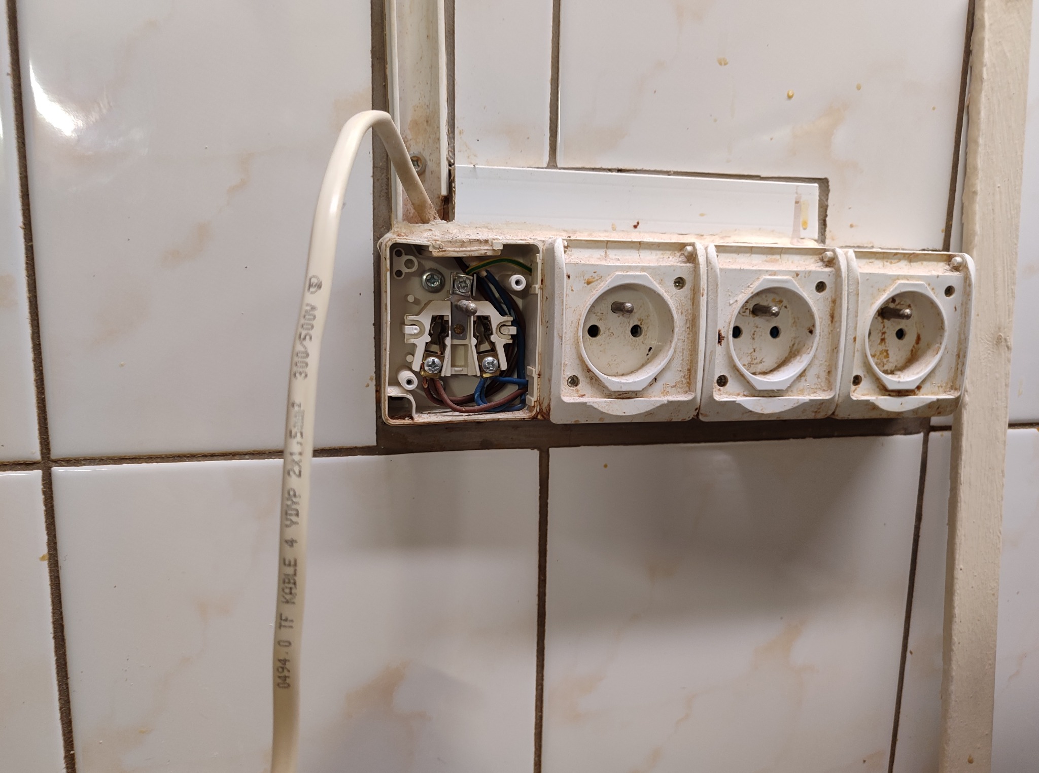 Otwarta puszka elektryczna z gniazdkami i wystającym przewodem na białej, brudnej ścianie w Szczecinie. Widoczne kable i instalacja elektryczna.