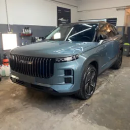 Błękitny SUV Jaecoo w warsztacie, widok z przodu, lśniąca karoseria, czyste felgi, oświetlenie warsztatowe, butelki z preparatami na półce w tle, plakat na ścianie.