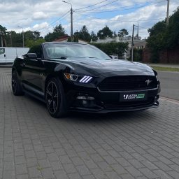 Perfect Clean Car detailing - Czarny kabriolet Ford Mustang z widocznym logo Ultra Coat Pro na przednim zderzaku, zaparkowany na szarej kostce brukowej w słoneczny dzień.