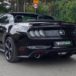 Perfect Clean Car detailing - Czarny kabriolet Ford Mustang GT z logo 'Ultra Coat Pro' na tylnej klapie, zaparkowany na kostce brukowej, z widocznymi znakami drogowymi i zielenią w tle.
