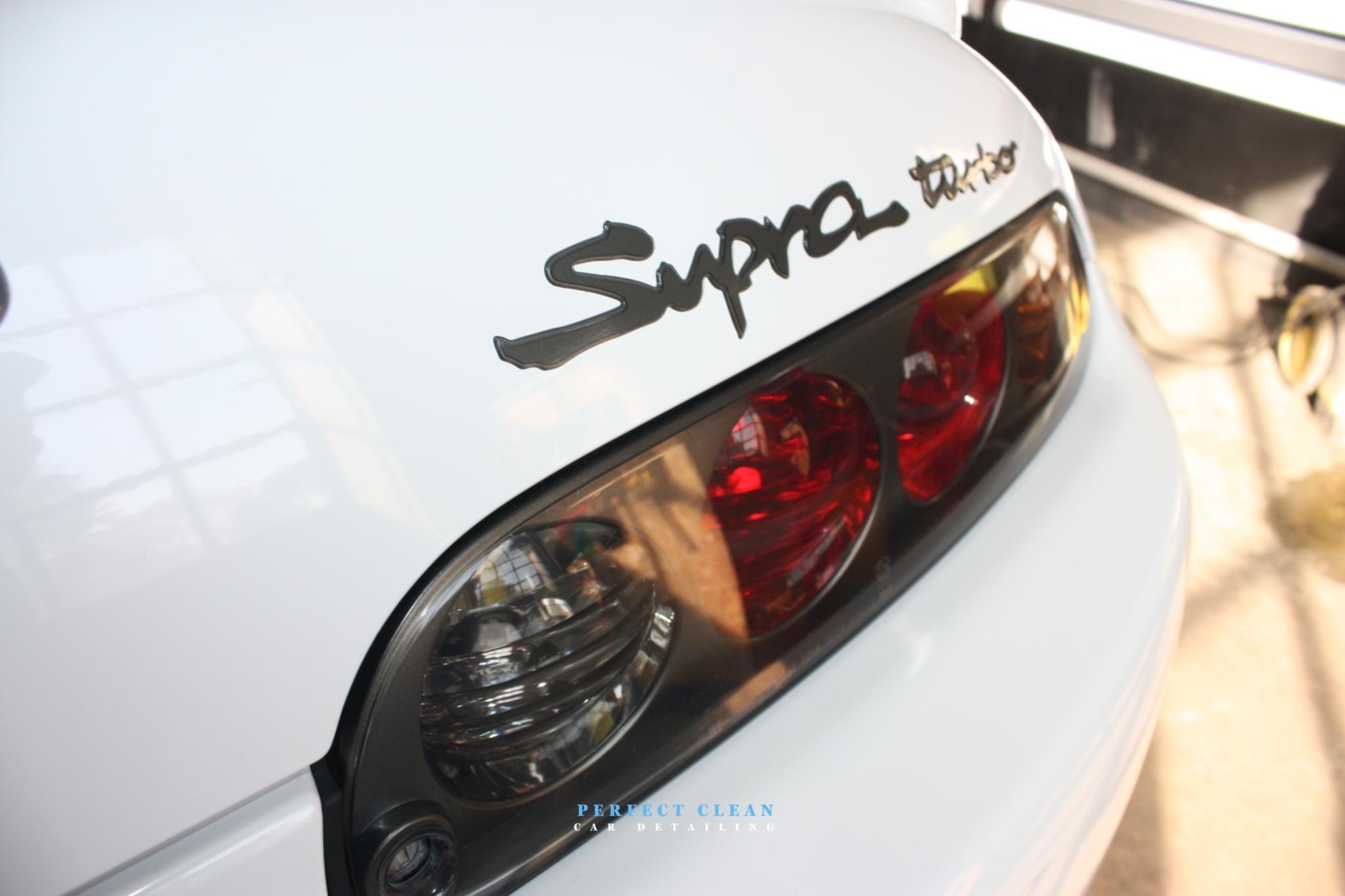 Biała Toyota Supra Turbo: zbliżenie na tylne światła i logo. Widoczne odbicia światła na lakierze, sugerujące świeże czyszczenie. Detailing.