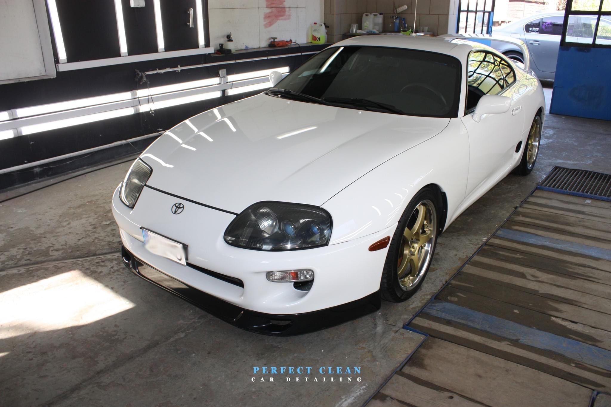 Biała Toyota Supra z złotymi felgami po detailingu, widoczna w jasnym garażu z oświetleniem LED, na drewnianej platformie myjącej.