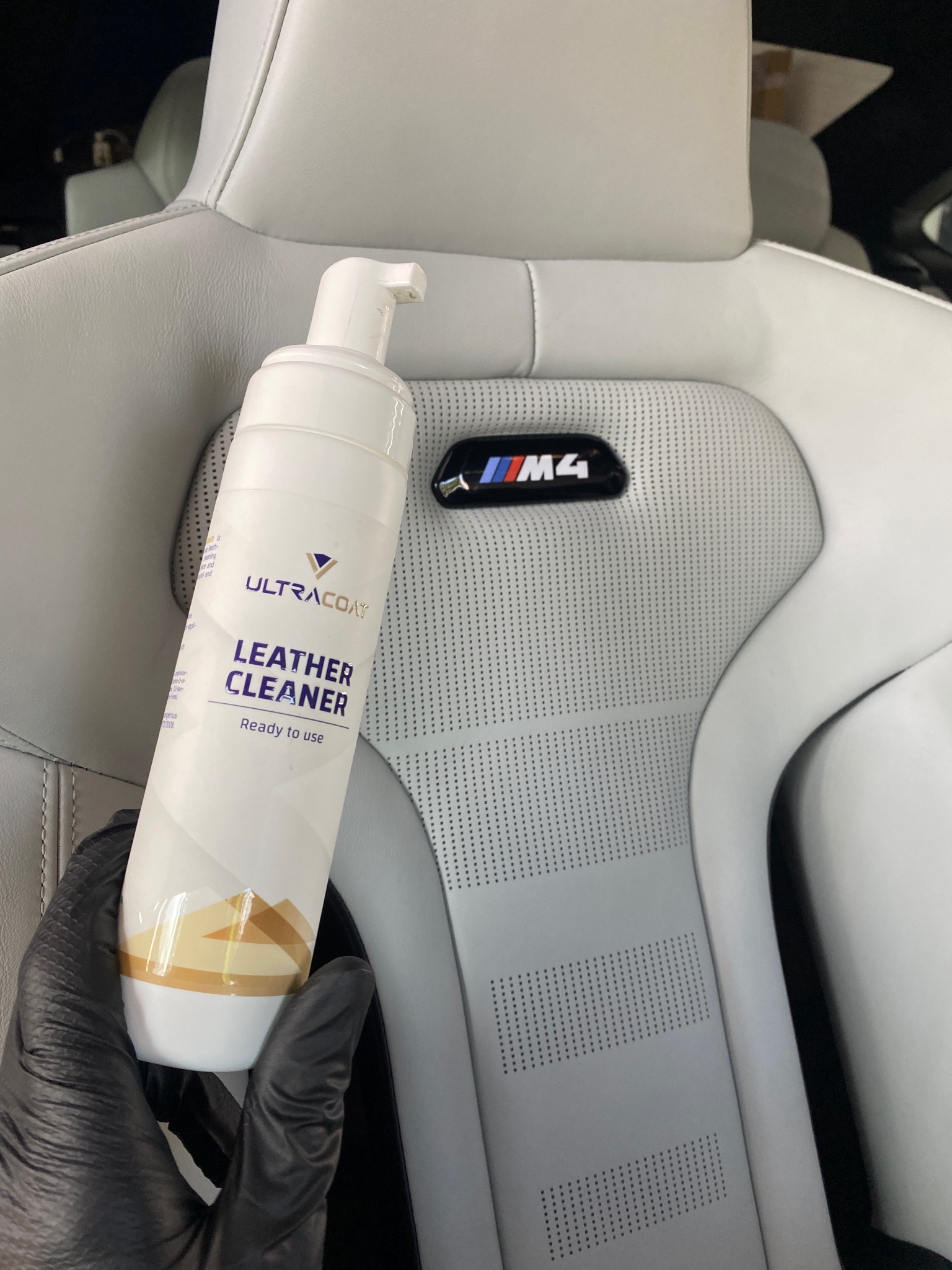 Białe skórzane siedzenie M4 z perforacją, czyszczone preparatem Ultracoat Leather Cleaner, trzymanym w dłoni w czarnej rękawicy. Detailing wnętrza.