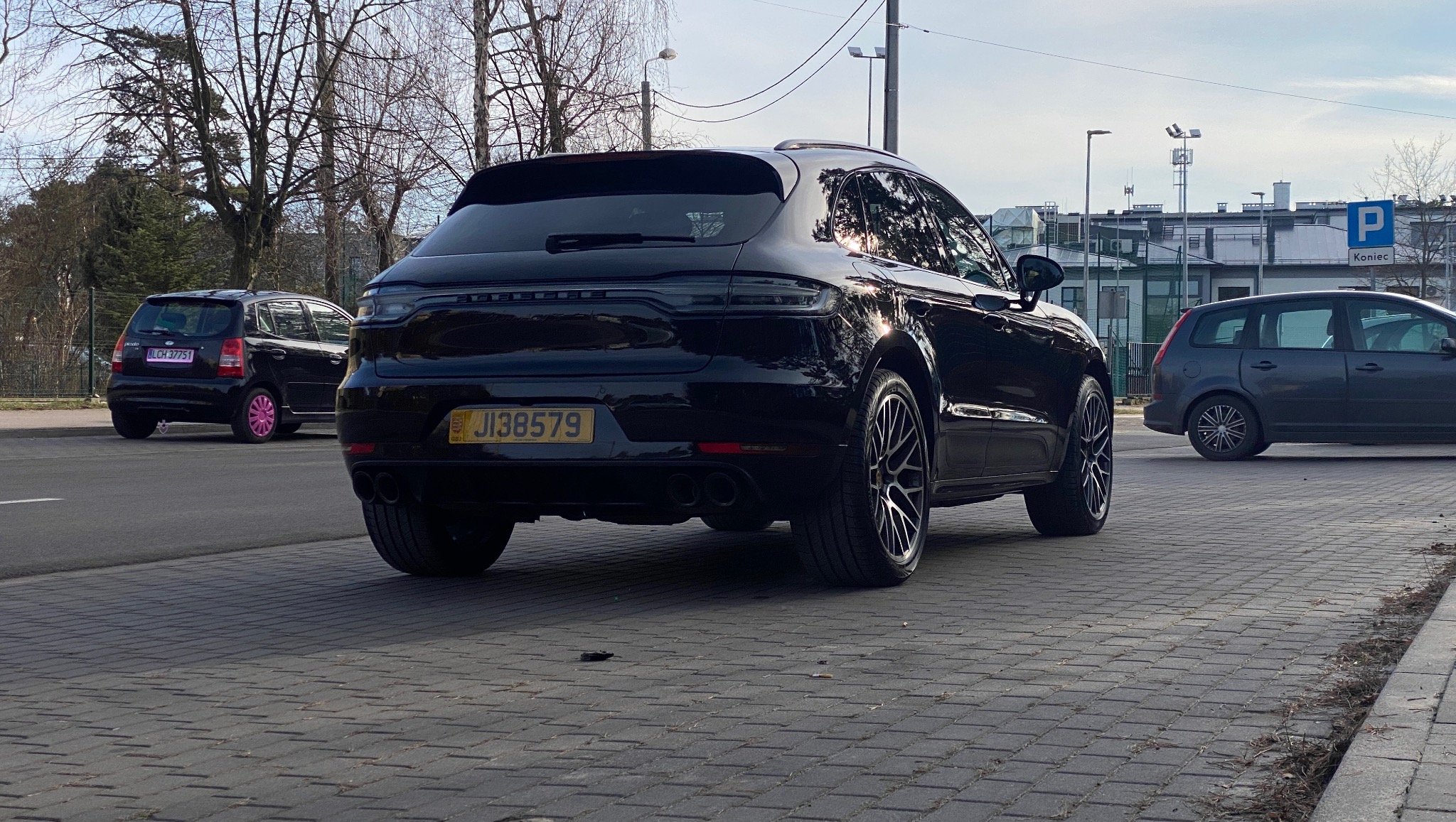 Czarny Porsche Macan z tablicą rejestracyjną JI38579, widok z tyłu na tle miejskiego krajobrazu z innymi autami i znakami drogowymi.