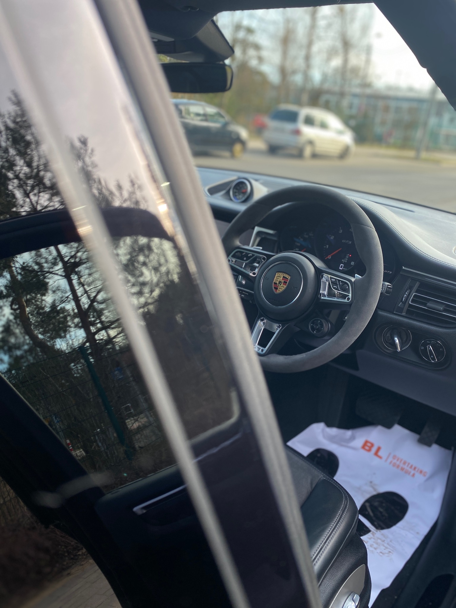 Wnętrze Porsche Macan GTS: kierownica obszyta alcantarą, widoczny kokpit i firmowa mata pod nogami. Detal z zewnątrz przez szybę.