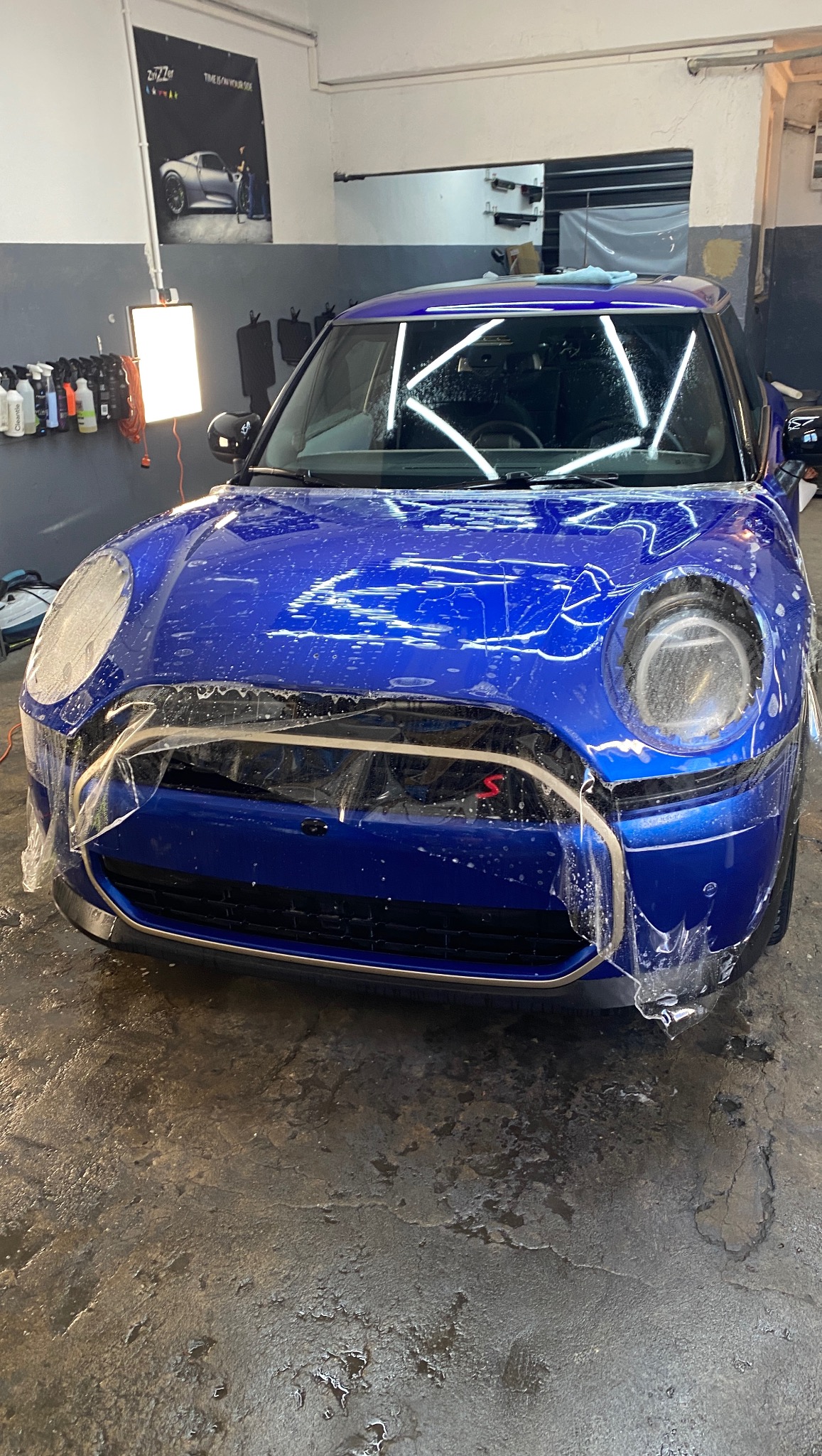 Niebieski Mini Cooper zabezpieczony folią ochronną w warsztacie, przygotowany do dalszych prac detailingowych, widoczne oświetlenie i plakat reklamowy.
