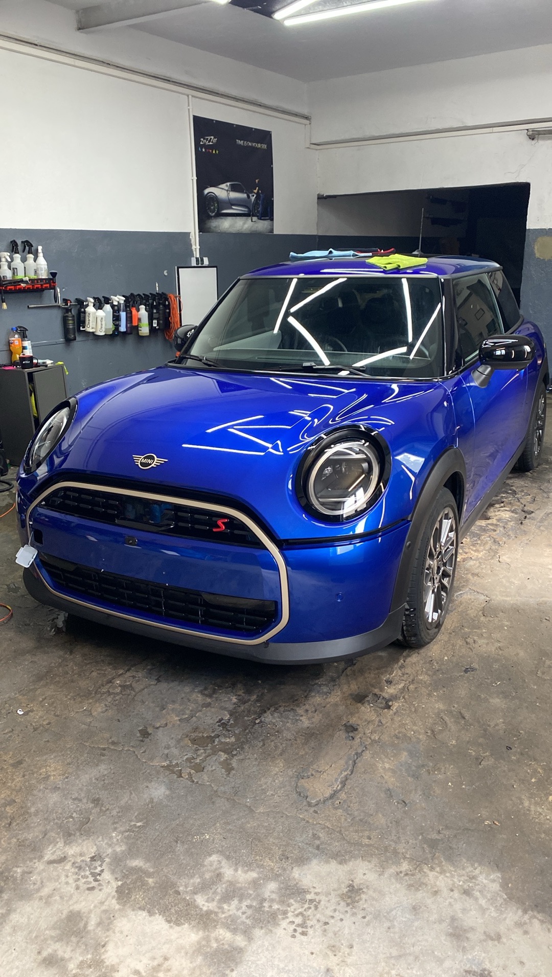 Błyszczący, niebieski Mini Cooper S po detailingu w warsztacie, widoczne odbicia światła, czysta karoseria, narzędzia i środki czystości w tle.