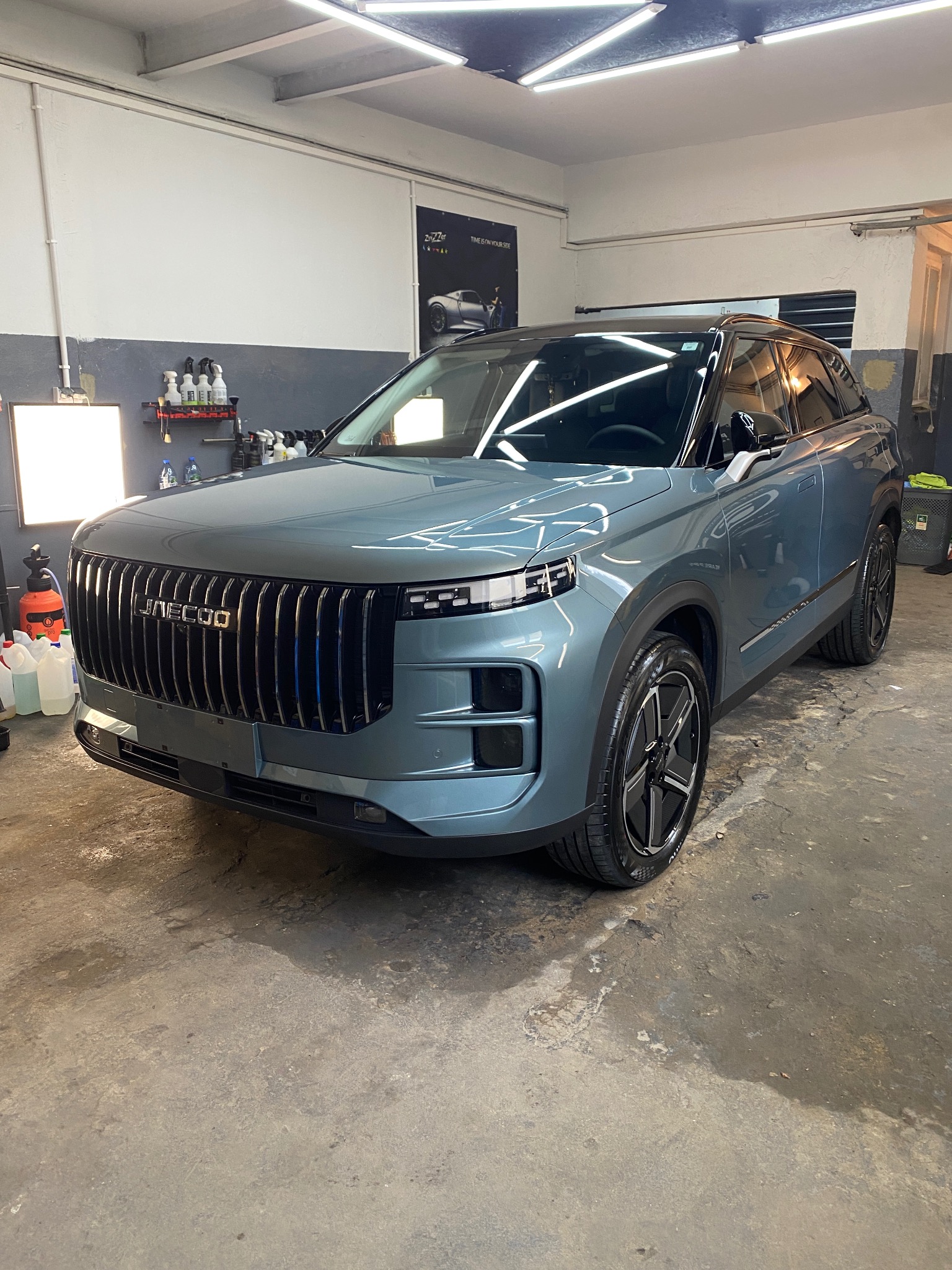 Błękitny SUV Jaecoo w warsztacie, widok z przodu, lśniąca karoseria, czyste felgi, oświetlenie warsztatowe, butelki z preparatami na półce w tle, plakat na ścianie.