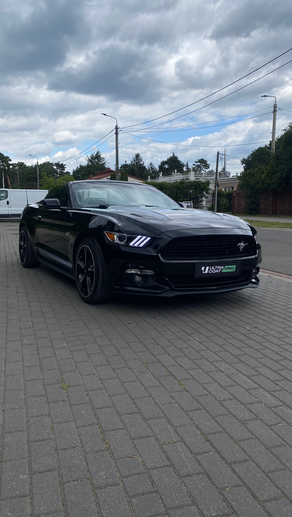 Czarny kabriolet Ford Mustang z widocznym logo Ultra Coat Pro na przednim zderzaku, zaparkowany na szarej kostce brukowej w słoneczny dzień.