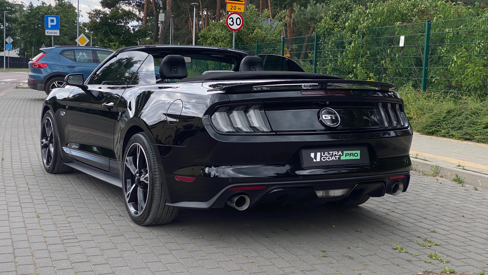 Czarny kabriolet Ford Mustang GT z logo 'Ultra Coat Pro' na tylnej klapie, zaparkowany na kostce brukowej, z widocznymi znakami drogowymi i zielenią w tle.
