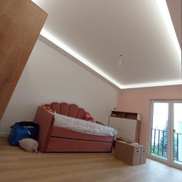 Dawid Rogalski - Pokój na poddaszu z oświetleniem LED na suficie, różową sofą i szafką. Widoczne kartony i worki. Podłoga drewniana. Świeże, jasne wnętrze po remoncie.