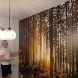 Dawid Rogalski - Mężczyzna prezentuje fototapetę z motywem leśnym, zamontowaną na ścianie obok drewnianych lameli. Widoczne lampy wiszące i okno z widokiem na zewnątrz.
