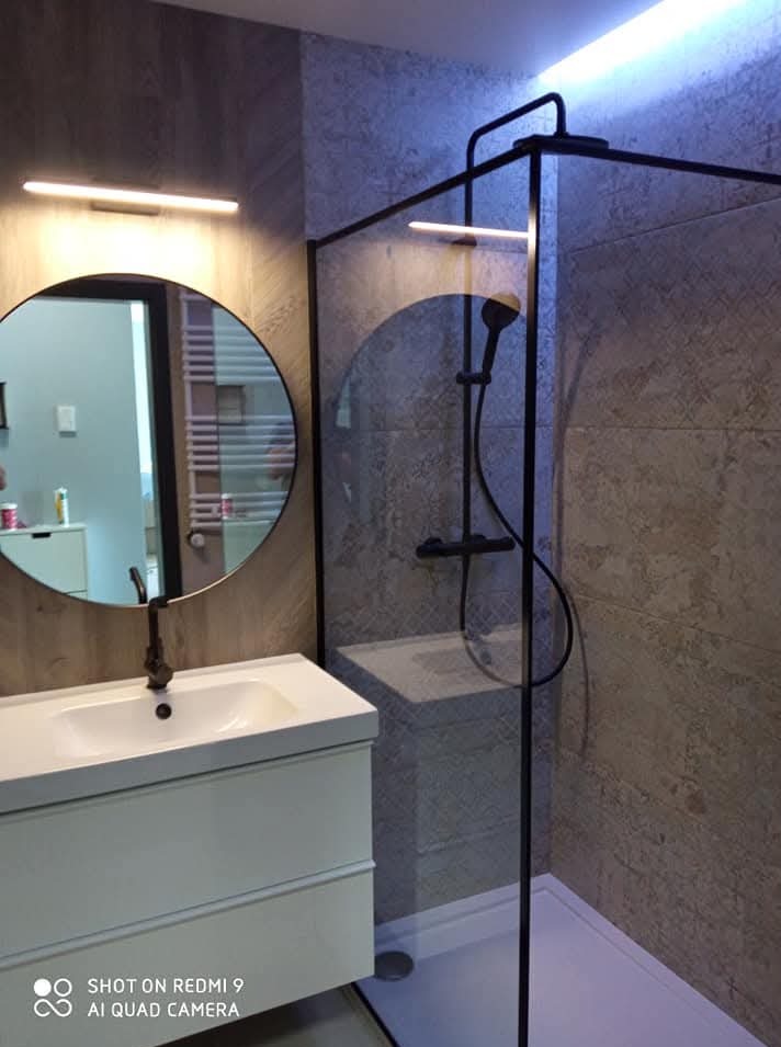 Nowoczesna łazienka z kabiną prysznicową typu walk-in, okrągłym lustrem i białą szafką pod umywalką. Stonowana kolorystyka, oświetlenie LED.