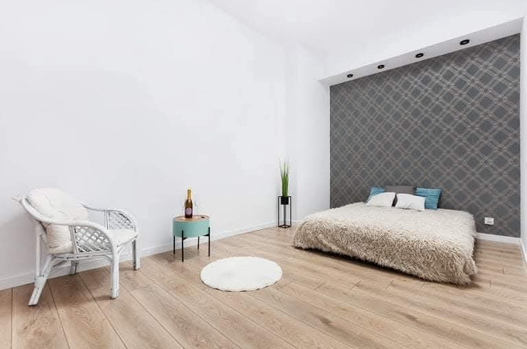 Jasne panele podłogowe w sypialni z szarą ścianą w romby. W tle łóżko z puszystą narzutą. Na pierwszym planie biały fotel rattanowy i okrągły dywanik.