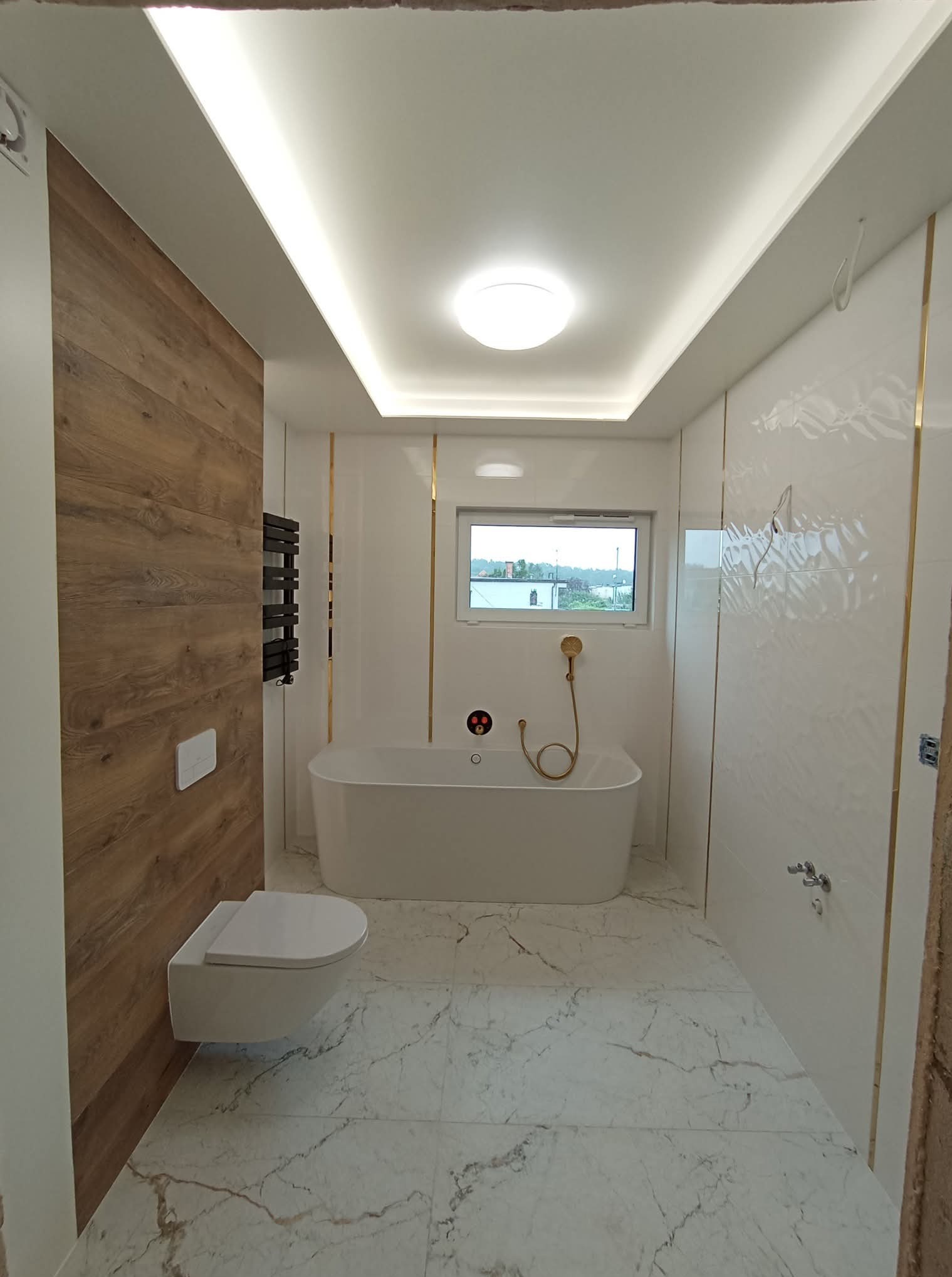 Elegancka łazienka z wanną wolnostojącą, drewnianą ścianą i marmurową podłogą. Białe płytki z złotymi akcentami. Nowoczesne oświetlenie sufitowe.