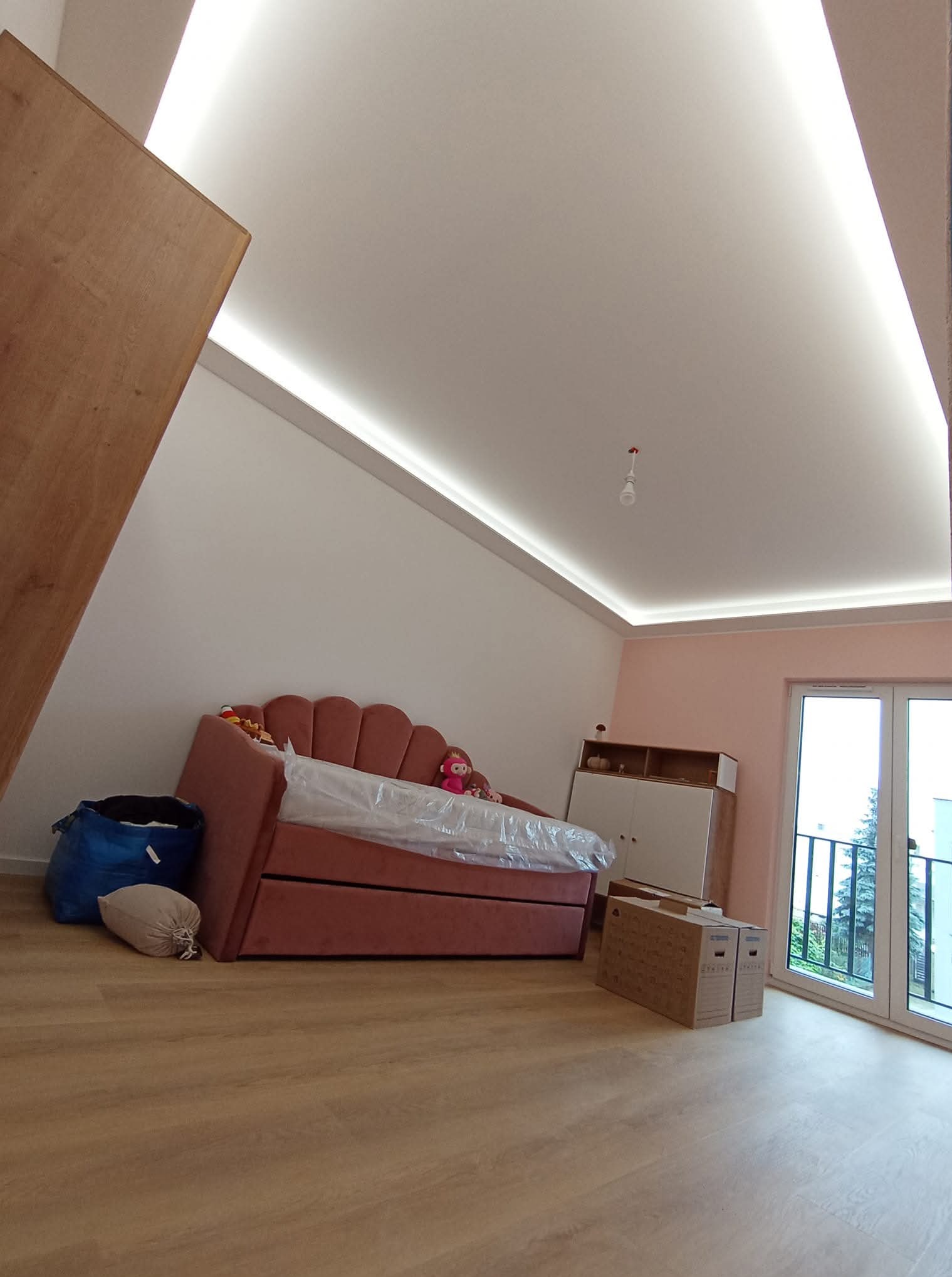 Pokój na poddaszu z oświetleniem LED na suficie, różową sofą i szafką. Widoczne kartony i worki. Podłoga drewniana. Świeże, jasne wnętrze po remoncie.