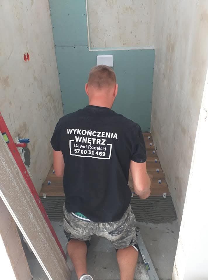 Glazurnik w trakcie pracy: układanie drewnopodobnych płytek w wąskiej łazience. Widoczna ściana z płyt karton-gips i narzędzia. Na koszulce logo firmy.