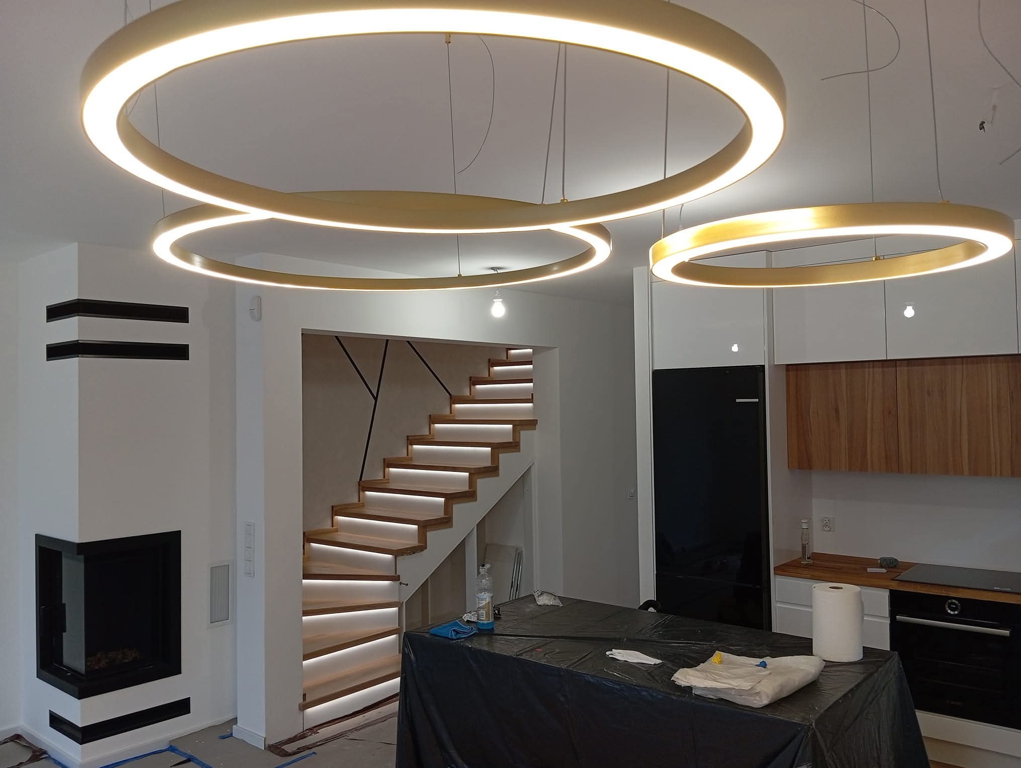 Nowoczesne wnętrze z podświetlanymi schodami i okrągłymi lampami. W tle widoczna kuchnia z drewnianymi elementami i czarną lodówką.