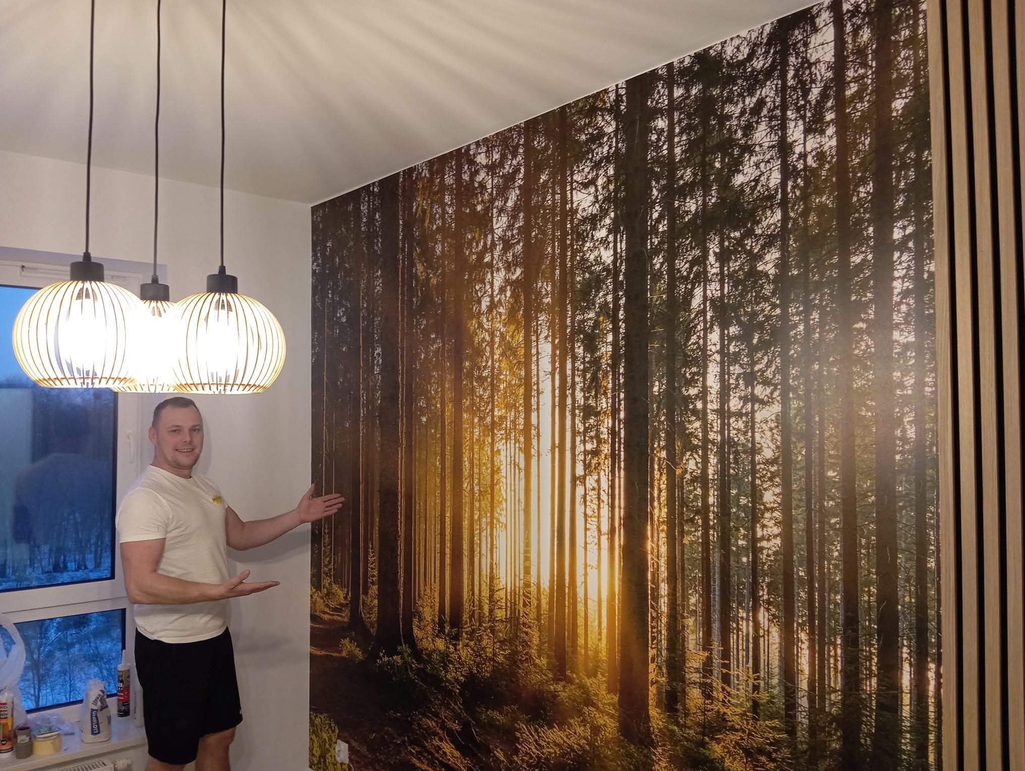 Mężczyzna prezentuje fototapetę z motywem leśnym, zamontowaną na ścianie obok drewnianych lameli. Widoczne lampy wiszące i okno z widokiem na zewnątrz.