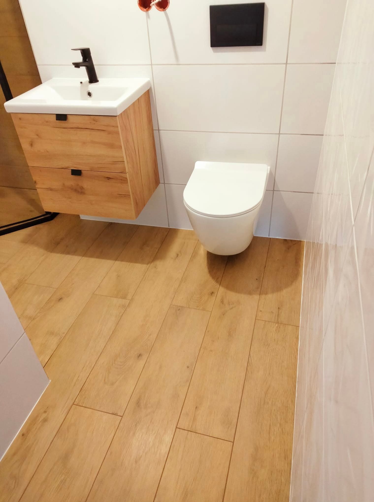 Nowoczesna łazienka po remoncie: drewnopodobna podłoga, biała ceramika, wisząca szafka z umywalką i czarna armatura. Minimalistyczny design, płytki na ścianach.